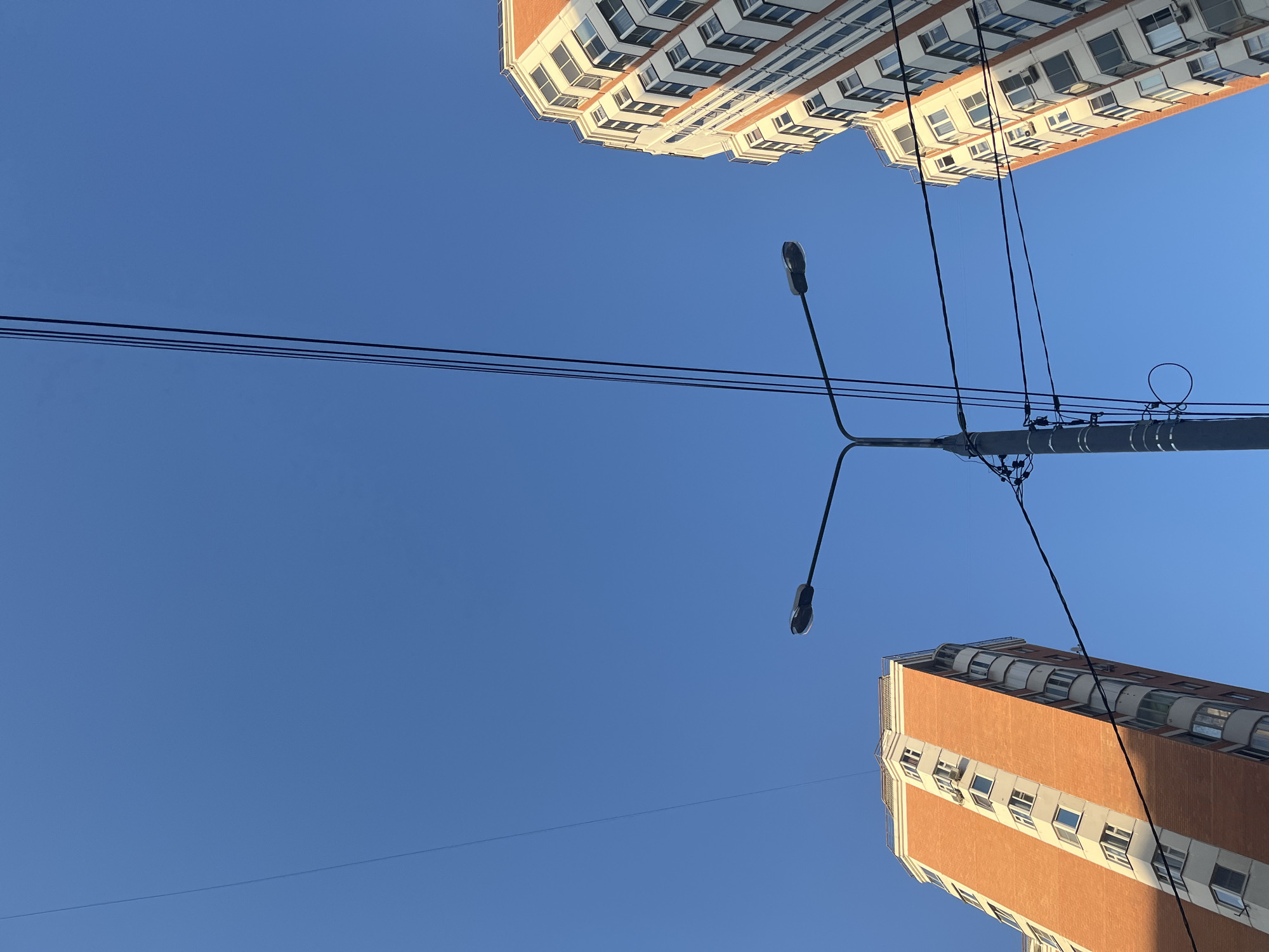 The image captures a low-angle view of an urban environment under a clear, cloudless blue sky. No people are visible.
Two multi-story buildings flank a centrally located dark gray street light pole. The building on the left, positioned towards the lower-left corner and extending upwards, features orange-red brick facades with numerous white-framed windows. Several air conditioning units are affixed to its visible side. The right face of this building is illuminated by sunlight, while its left face and some window recesses are in shadow. A rounded, multi-windowed section is visible along its right edge.
The building on the right, extending from the lower-right corner upwards, displays a similar architectural style with orange-red and white sections. It features prominent, multi-faceted bay windows or rounded balcony structures running vertically along its visible facade. This building is also brightly illuminated by direct sunlight, casting minimal shadows on its immediate surface.
The central street light pole is dark gray or black, featuring a T-shaped cross arm at its top supporting two luminaires, one oriented to the left and one to the right. The pole itself is densely wrapped with various dark cables, wires, and attachment hardware. A distinct coiled loop of black cable is visible near the base of the pole's visible section.
Numerous black electrical or communication wires crisscross the expansive blue sky. A single, thin wire runs diagonally from the upper-left quadrant towards the lower-middle. A parallel group of three distinct, slightly thicker wires runs diagonally from the upper-middle section of the sky towards the lower-right. Several other, thicker, intertwined wires are affixed to the street light pole and extend horizontally and diagonally across the mid-ground, passing in front of the buildings. One particularly thick, dark cable runs from the pole towards the lower-right, passing in front of the lower section of the left building.
The strong sunlight and clear sky suggest a bright daytime setting. The location is Unknown, Unknown.