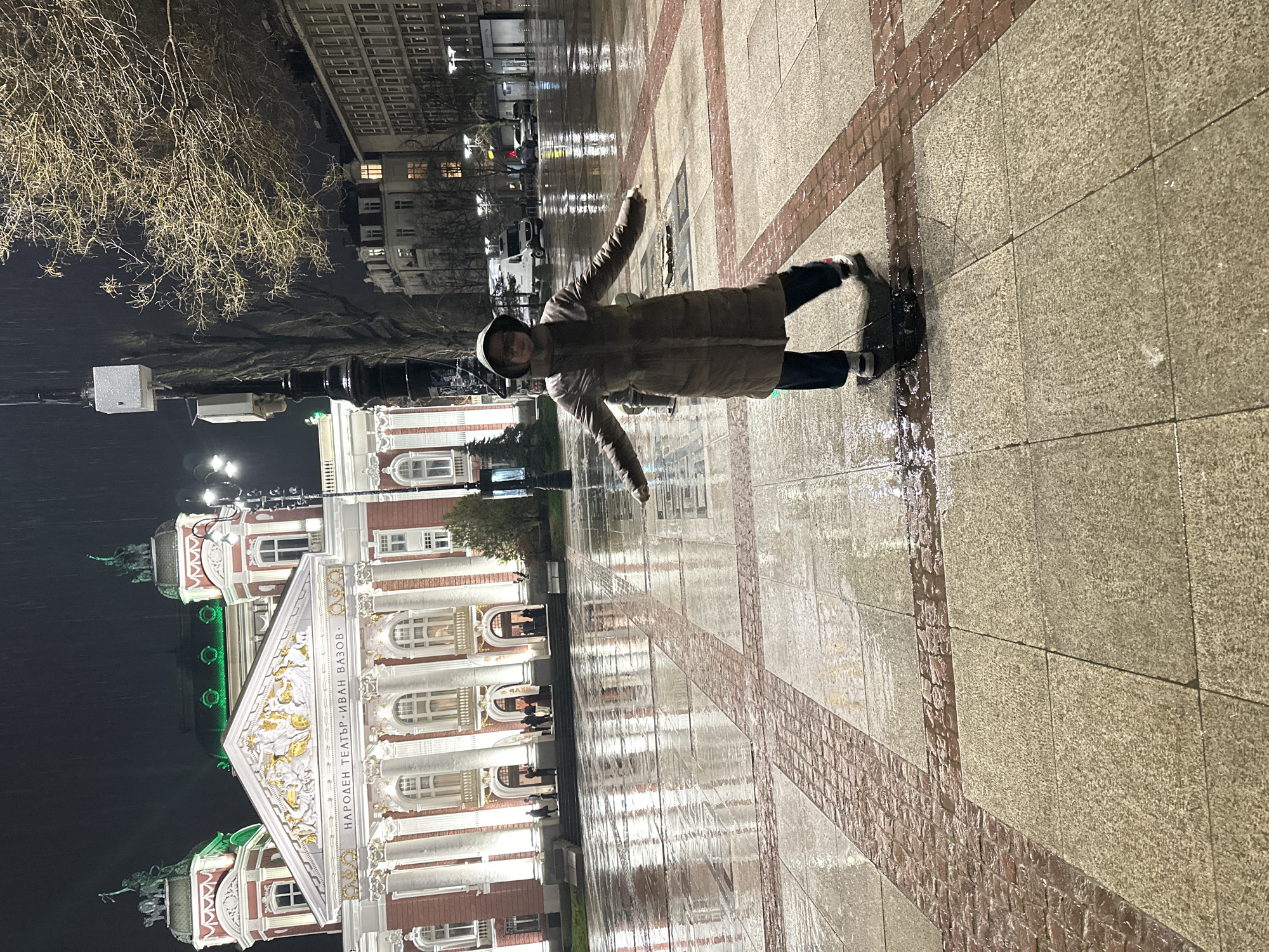 A nighttime photograph depicts an individual posing on a wet paved plaza in front of the Ivan Vazov National Theatre in Sofia, Bulgaria. The central figure, a person with short hair, wears a hooded, light brown, knee-length puffer coat, dark trousers, and light-colored sneakers, standing with arms horizontally outstretched. The wet, grey and red-bricked pavement reflects the bright artificial lights illuminating the scene. In the background, the Ivan Vazov National Theatre, a classical red and white building, is prominently illuminated, featuring columns, a golden pediment with relief sculptures, and two towers with green-lit domes and statues. The theatre's facade displays the Cyrillic text "НАРОДЕН ТЕАТЪР „ИВАН ВАЗОВ“". Several indistinct figures are visible on the theatre's entrance steps. To the right of the central figure, a black multi-headed streetlight pole with attached white utility boxes stands next to a large, bare-branched tree. Beyond, other urban buildings with illuminated windows and a street with vehicles are visible. Faint streaking in the upper portion of the image suggests ongoing light rain.
