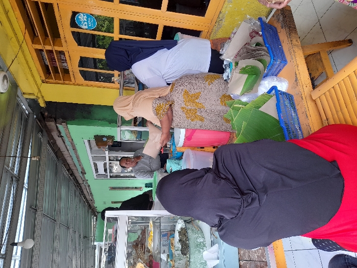 Three women stand at a table outdoors, one in a blue hijab and white shirt, another in a tan hijab and patterned dress, and the third in a black hijab and red shirt. They appear to be preparing food, surrounded by green leaves and blue plastic baskets. A man sits inside a nearby window, and a display case sits in front of the women. The outdoor area features a yellow and green building with a metal awning.