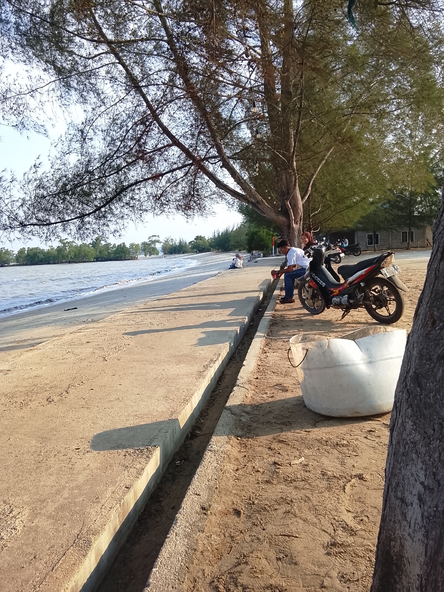 The image depicts a scene by the water in Lubuk Puding, Indonesia. A concrete embankment runs along a sandy beach, with the sea visible to the left. Several people are present. In the foreground, a motorcycle is parked on the sand next to a large white bucket. Two individuals, a man and a woman, are seated on the embankment near the motorcycle. The man is wearing a white shirt and blue jeans, holding a cup. The woman, wearing a hijab, is seated next to him. Further down the embankment, another person is seated on the sand, and in the distance, more people are visible near the water's edge. Trees line the background, with buildings visible behind them. The sun is casting long shadows, indicating it may be late afternoon or early morning.