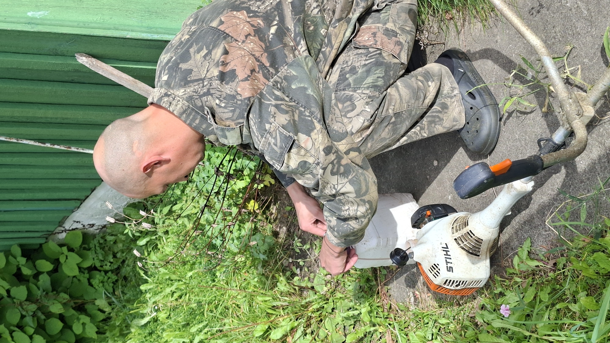 A bald, fair-skinned man wearing camouflage overalls and black Crocs crouches on a paved surface next to a green wooden fence. He is holding a white plastic jug with his right hand. To his left, a white and orange string trimmer with the STIHL logo lies on the ground. The scene appears to be outdoors, with green foliage and a rough concrete patch visible. The man's attention is directed towards the jug. The environment suggests a rural or suburban setting, possibly a garden or yard.