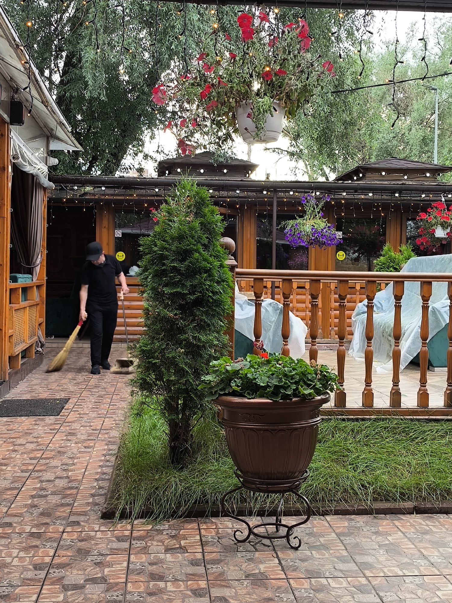 The image depicts an outdoor scene at a restaurant with wooden structures and greenery. A person wearing a black t-shirt, black pants, and a black cap is sweeping the tiled walkway with a broom. The person is positioned to the left of a tall, conical green shrub. In the foreground, to the right, is a large brown planter with green foliage, supported by an ornate black metal stand. Behind the shrub, there is a wooden railing with vertical posts. Beyond the railing, there are chairs covered with light-colored plastic sheeting. The background features a wooden building with windows, adorned with strings of warm-toned fairy lights. Hanging baskets with red and purple flowers are suspended from above, and large trees with green foliage are visible in the distance. The overall setting appears to be a patio or outdoor seating area of a restaurant.