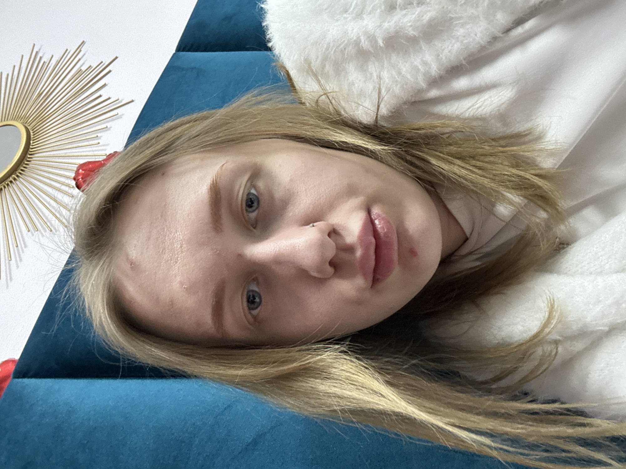 A close-up, rotated view of a young woman with light blonde hair and fair skin. Her head is angled towards the right. She has light blue eyes and a nose piercing. She wears a white top with what appears to be a fluffy white collar or layer. The background includes a portion of a gold sunburst-shaped mirror and a blue, possibly velvet, surface.
There is no location information in the image, so I cannot provide its location in Rostov-on-Don, Russia.