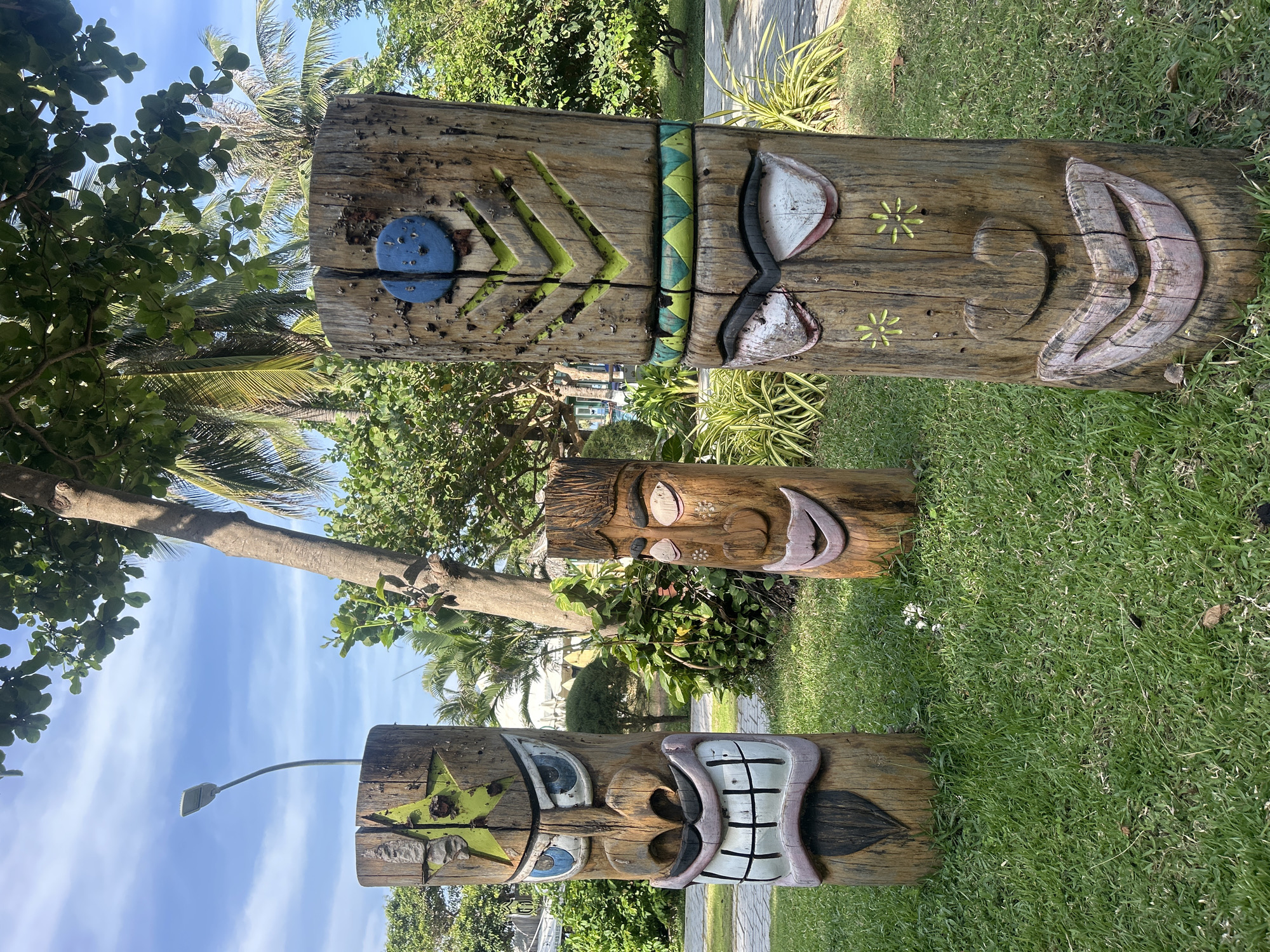 Three totem poles stand in a grassy area beneath trees, under a blue sky. The poles feature carved faces with colorful details. A streetlight is visible behind the closest pole. The location is likely Nha Trang, Vietnam, suggested by the presence of palm trees and tropical foliage. No people are visible in the scene.