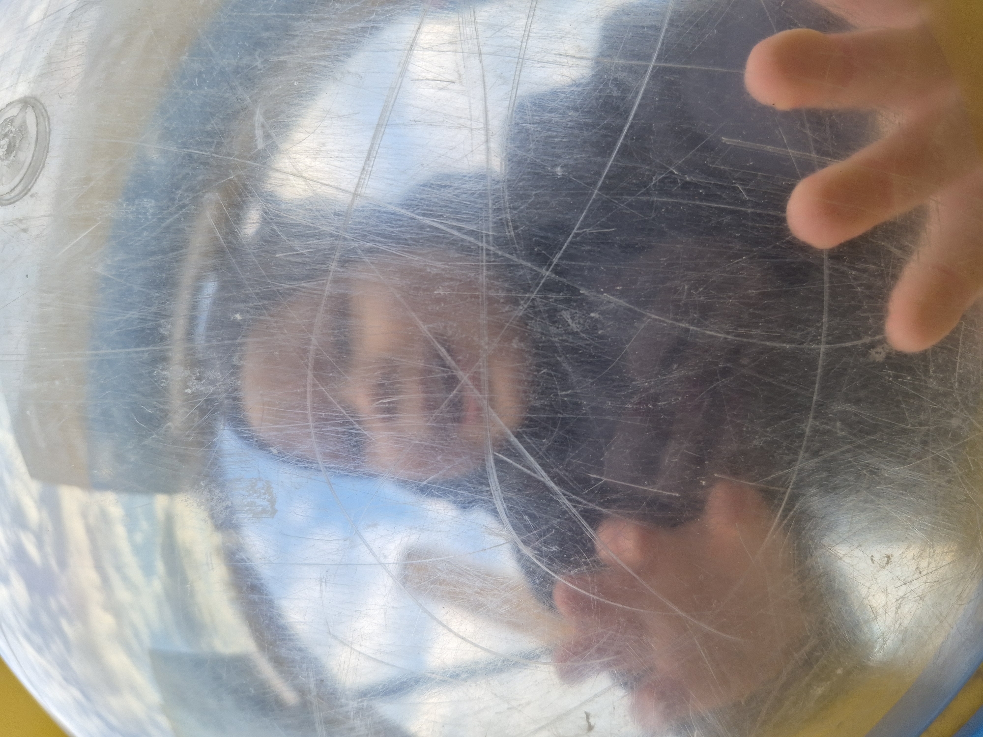 The image shows a blurred view through a scratched transparent surface, possibly a dome or bubble-like structure. A child's face and hands are visible, pressed against the surface from the inside. The background appears to be outdoor with hints of sky and possibly other structures. There is no information in the image to suggest a location in Tobolsk, Russia.