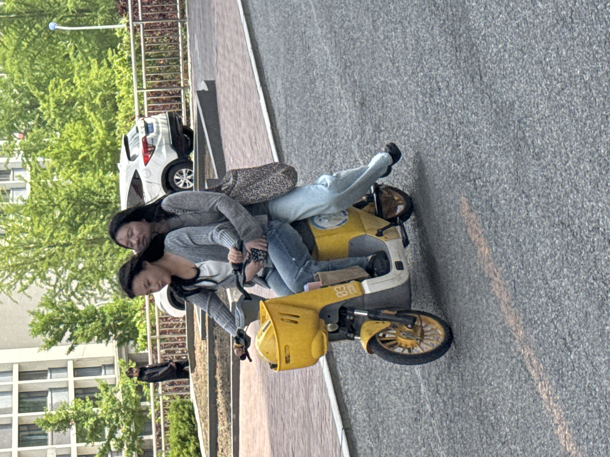 Two women are riding a yellow moped on a paved surface next to a sidewalk. The woman in the front is driving, wearing a gray sweater and jeans. The woman behind her is leaning against her back, wearing a similar gray sweater and jeans. Behind them is a white car parked next to a metal fence. Further back, there are trees and buildings. The image appears to have been taken in Zhongtun, China, as indicated by the prompt. The overall environment seems to be an urban street scene.