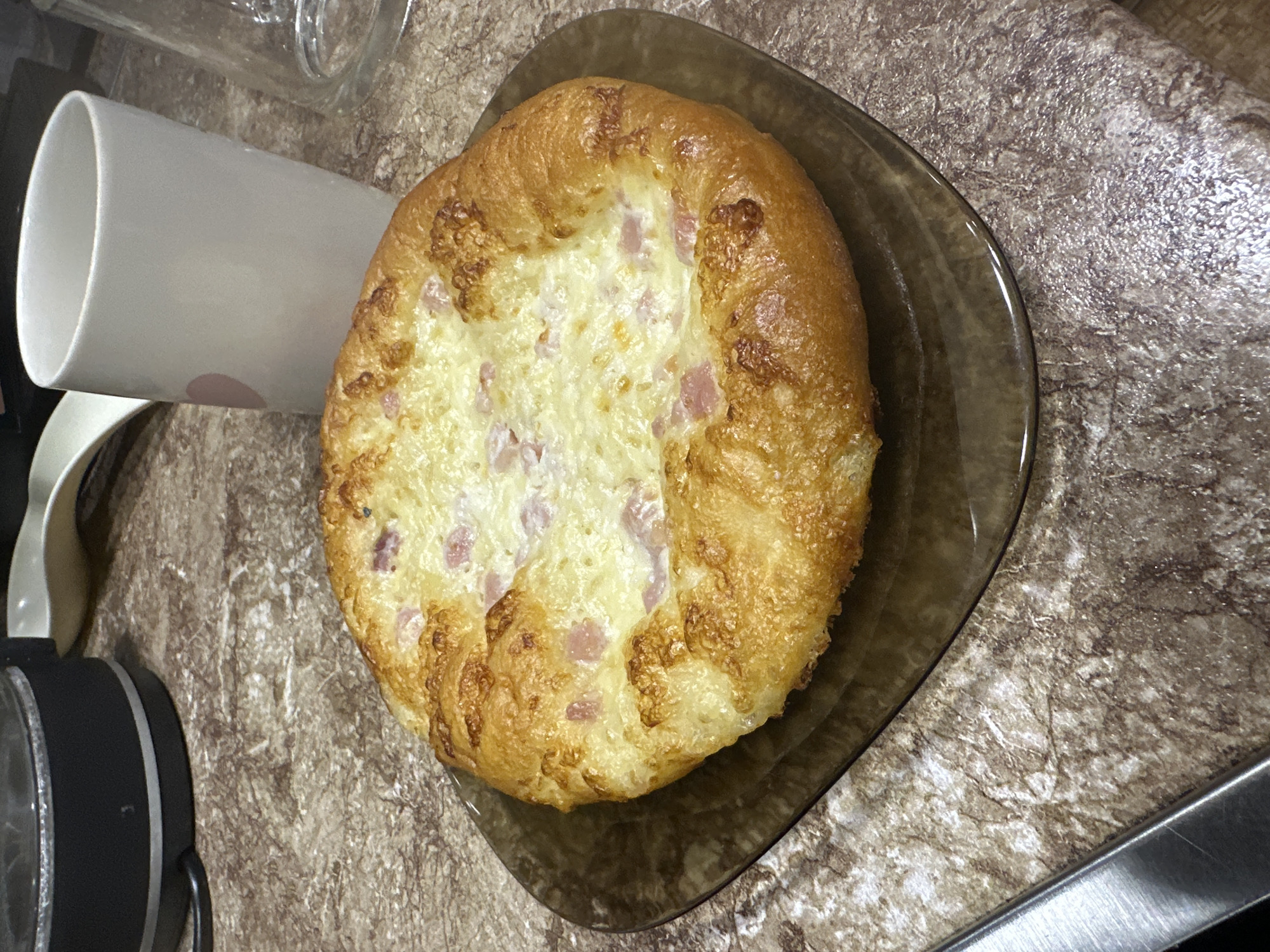 The image shows a close-up, top-down view of a single, savory pastry, likely a type of bun or bread, served on a dark brown, translucent plate. The pastry has a golden-brown, slightly puffed crust that is slightly irregular in shape. The center of the pastry is filled with a creamy, white mixture speckled with small pink pieces, which appear to be diced ham or a similar cured meat, and topped with melted cheese that has browned in places.
To the left of the pastry, a portion of a white mug with a light pink circular design is visible, tilted on its side. Further to the left, a black and silver object that resembles headphones is partially visible at the bottom edge of the frame. The background is a speckled, mottled surface that resembles a countertop or table, with a greyish-brown and white pattern.
The lighting appears to be artificial, casting some subtle shadows, and the scene suggests an indoor setting, possibly a kitchen or dining area. There is no clear indication of the time of day or weather. The focus is on the food item, giving it a prominent and appetizing appearance. The overall impression is of a prepared meal or snack. No visible text is present in the image.