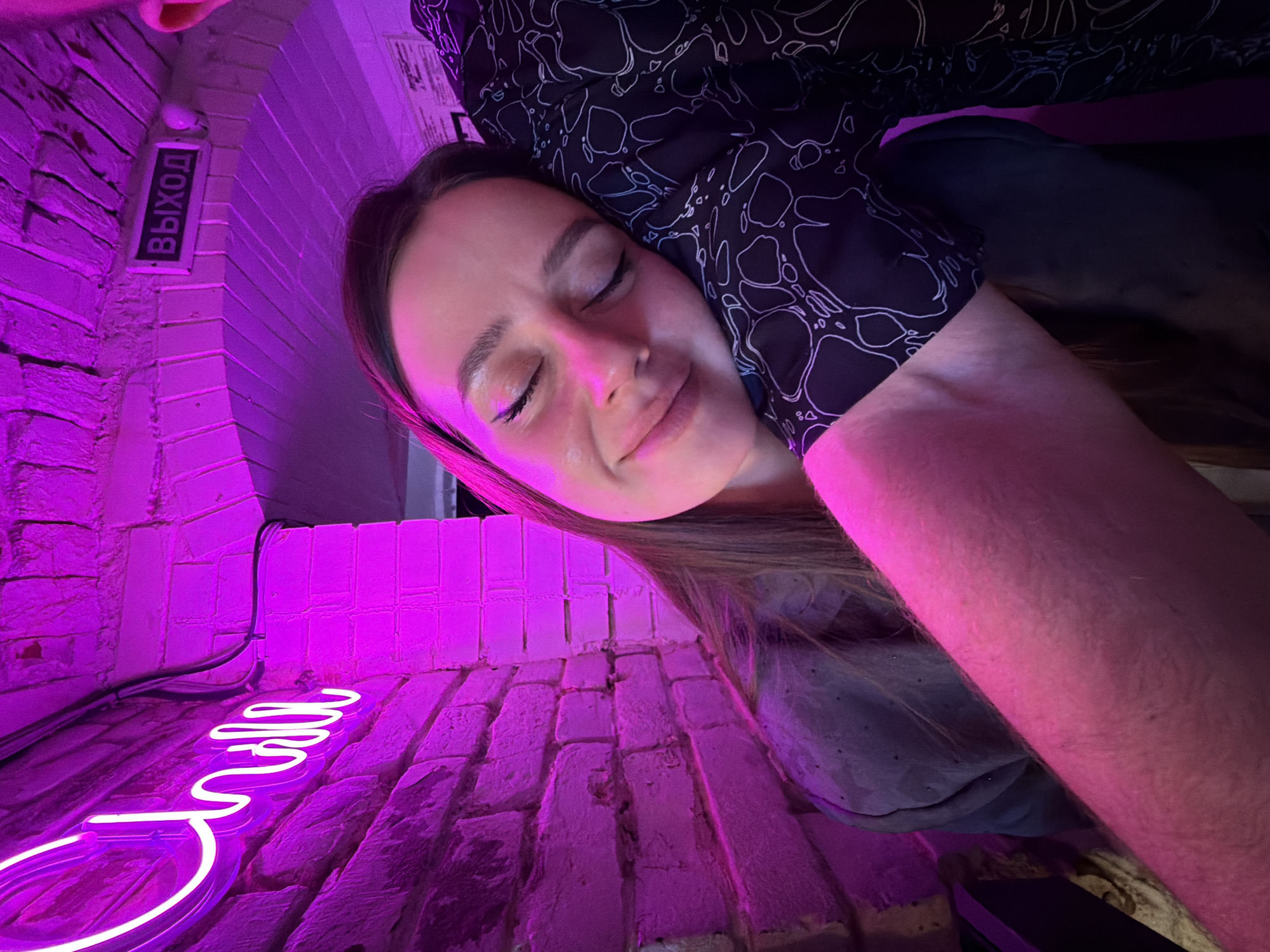 This close-up, tilted image features a young woman resting her head against what appears to be another person's arm or shoulder. Her eyes are closed, and a peaceful, faint smile graces her lips, conveying a sense of relaxation or contentment. Her dark hair frames her face, which is prominently illuminated by a vivid magenta-purple light.
The setting appears to be indoors, likely a bar, club, or cafe, characterized by brick walls and artificial lighting. These brick surfaces, which serve as both background and what seems to be the floor due to the image's rotation, are intensely saturated with the purple glow. In the upper left, an illuminated sign clearly displays the Cyrillic word "ВЫХОД" (Vykhod), meaning "EXIT" in Russian. In the lower left, a curved, white neon sign with stylized lettering is partially visible, though the complete word is indistinct. Some dark electrical cables are also visible running along the brickwork.
The atmosphere is intimate and moody, primarily due to the strong purple neon lighting, suggesting an evening or nighttime setting. The woman's serene expression indicates a moment of quiet repose. The presence of the "ВЫХОД" sign confirms a Russian-speaking environment, aligning with the specified location context of Krasnoyarsk, Russia.
