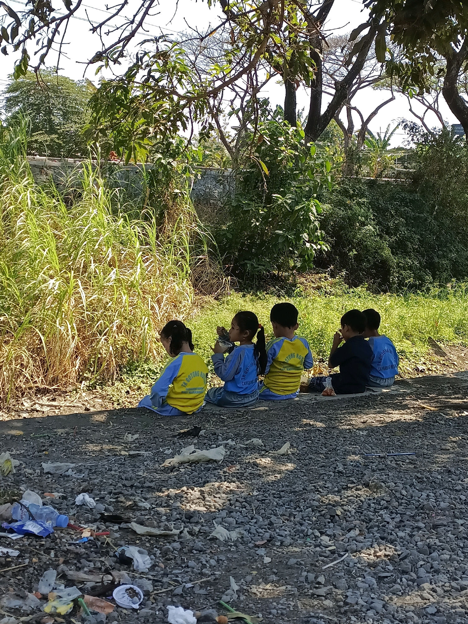 Five young children, presumably students, are sitting on a gravel path under trees. They are facing away from the camera, toward a dense area of tall, dry grass. The children are wearing school uniforms. The child on the far left wears a blue shirt and gray pants. The next child to the right wears a yellow shirt with blue sleeves and yellow pants. The child in the center wears a dark blue shirt and dark pants, and appears to be eating. To the right of this child, another child wears a blue shirt and gray pants. The child on the far right also wears a blue shirt and gray pants. Behind the children, a stone wall is visible, partially covered by vegetation. The area is littered with trash, including plastic bottles, wrappers, and various other debris. The scene appears to be outdoors, likely in a rural or semi-rural setting within Cangkring Rembang, Indonesia, given the presence of specific school uniforms. The time of day appears to be daytime, with strong sunlight casting shadows.