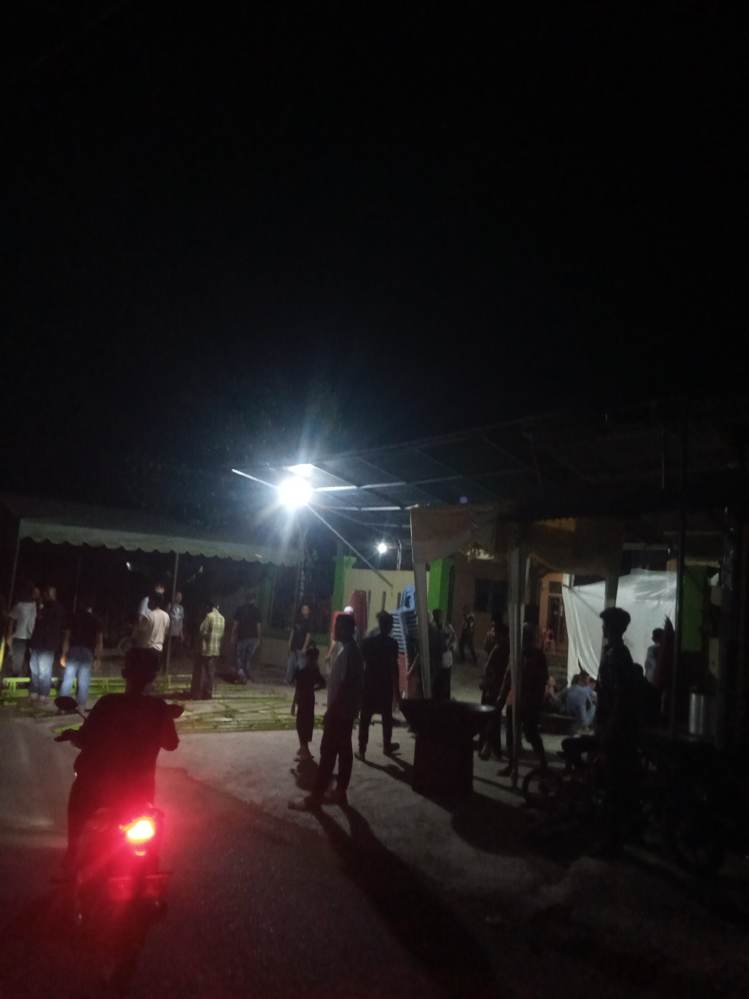 The image depicts a nighttime scene in the city of Paya Linteung, Indonesia, possibly at an outdoor gathering or event. A bright, artificial light source illuminates the area from above, creating strong shadows. Several individuals are visible, some standing and conversing, while others appear to be walking or observing. In the foreground, a person is riding a motorcycle with its red taillight brightly lit. Tents or awnings are set up, suggesting a temporary structure for the event. The environment appears to be a street or open area with some vegetation and buildings in the background. The overall atmosphere is dimly lit and appears to be a community gathering.