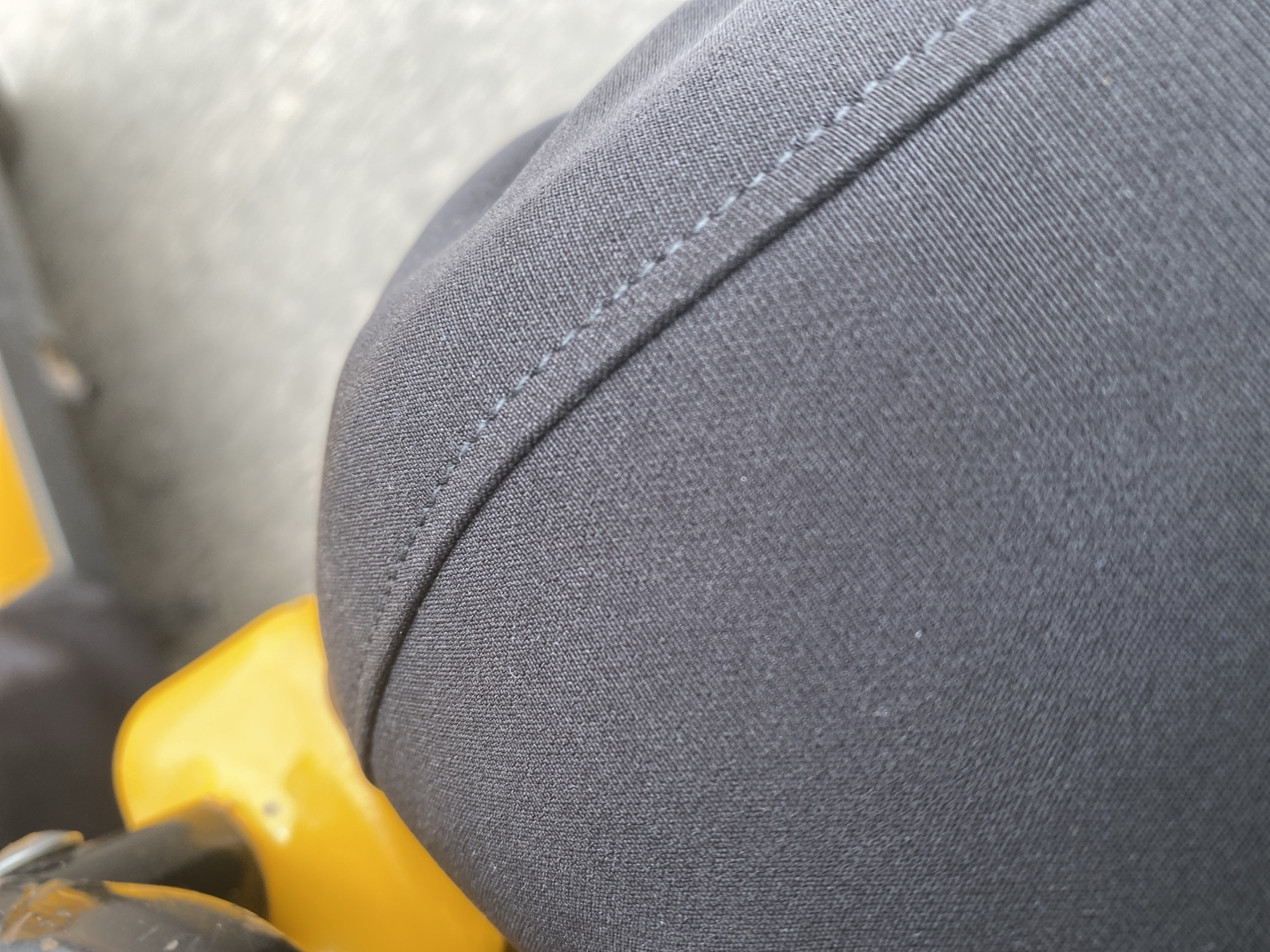 This is a close-up, high-angle shot of dark gray fabric with a visible seam. The fabric appears to be part of clothing, possibly pants or a skirt, and features a subtle, light blue thread for stitching along a curved edge. To the left of the fabric, a bright yellow object is partially visible, appearing to be a piece of plastic or painted metal. This yellow object has a curved shape and a smooth, glossy surface. The background is blurred and appears to be a neutral, light-colored surface, possibly concrete or asphalt, suggesting an outdoor setting. The lighting is bright, indicating daylight. There is no visible text or indication of specific time of day beyond daylight hours. The overall impression is that of a detailed texture study focusing on the fabric and its contrast with the yellow object, with no clear indication of people or a specific activity. The context of Jalingo, Nigeria, does not provide any direct visual cues within the image itself.