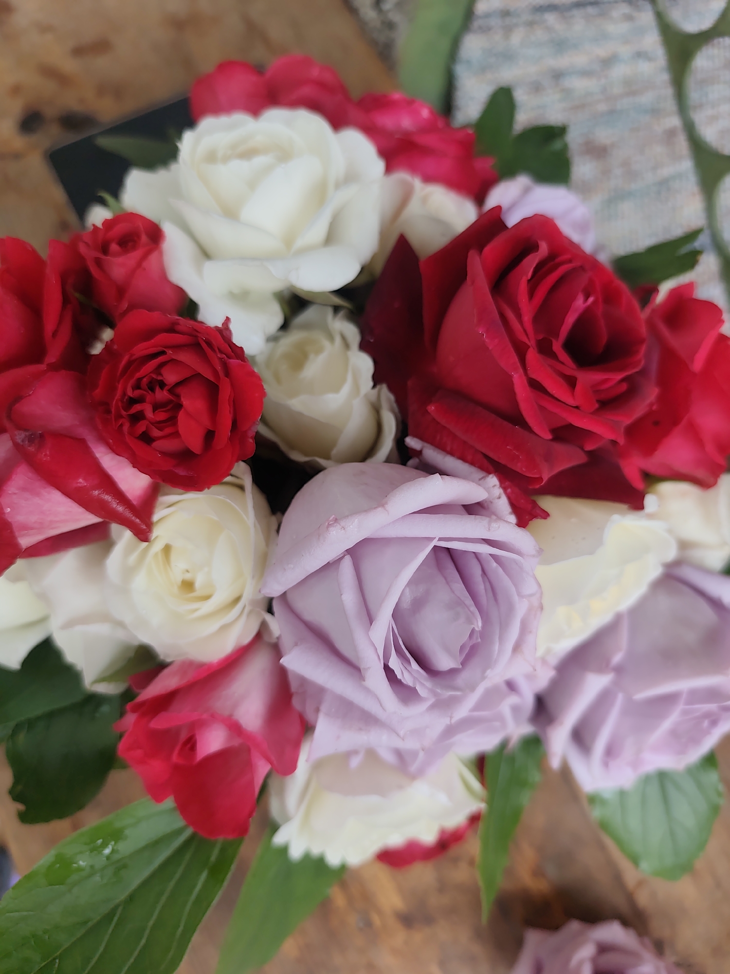 A close-up shot shows a bouquet of roses in various colors, including red, white, and lavender. The roses are arranged tightly together, creating a full and vibrant floral display. Several green leaves are interspersed among the blooms, providing a contrast to the colorful petals. The arrangement rests on a wooden surface. There is a black object on the upper left portion of the image. The city location is unknown.