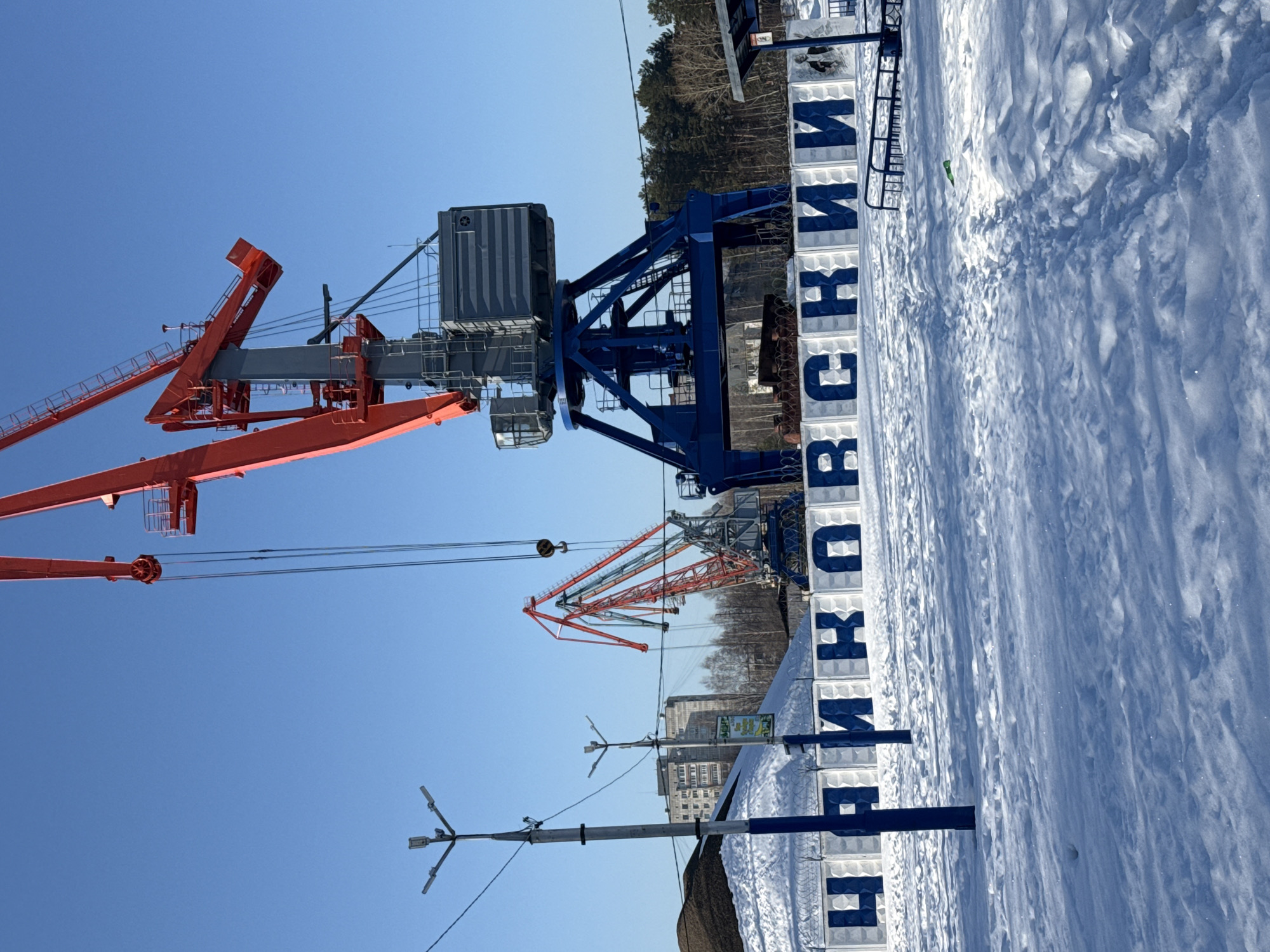 The scene depicts a snow-covered industrial or port area in the city of Chaikovsky, Russia, under a clear, bright blue sky. In the immediate foreground, the ground is entirely covered in undisturbed snow, crisscrossed by numerous footprints indicating recent pedestrian activity. A faint green object, possibly litter, is partially buried in the snow on the right.
Dominating the mid-ground is a long row of white concrete blocks forming a sign with the Cyrillic letters "ЧАЙКОВСКИЙ" (Chaikovsky) painted in bold blue. The concrete blocks are topped with coils of barbed wire. The final concrete block on the far right features a black and white depiction of a man's face.
Two large port cranes are prominent. A massive crane, located centrally and to the right, features a robust blue gantry-style base, a grey central tower, and a bright orange boom and arms extending upwards. Its operator's cabin is visible on the grey tower. To the left of this primary crane, another, smaller crane stands, primarily red with grey components.
To the far left, partially snow-covered piles of dark, granular material are visible. Two blue utility poles, each topped with a triple-arm street light fixture, stand in the left-midground. A small green sign is affixed to the second utility pole.
In the background, a line of bare deciduous trees and dark coniferous trees forms a horizon line. Beyond the trees, several multi-story residential buildings are visible in the distance. To the far right, next to the sign, is a black metal bench-like structure and a small shelter with a dark roof. No individuals are present or actively engaged in the scene.