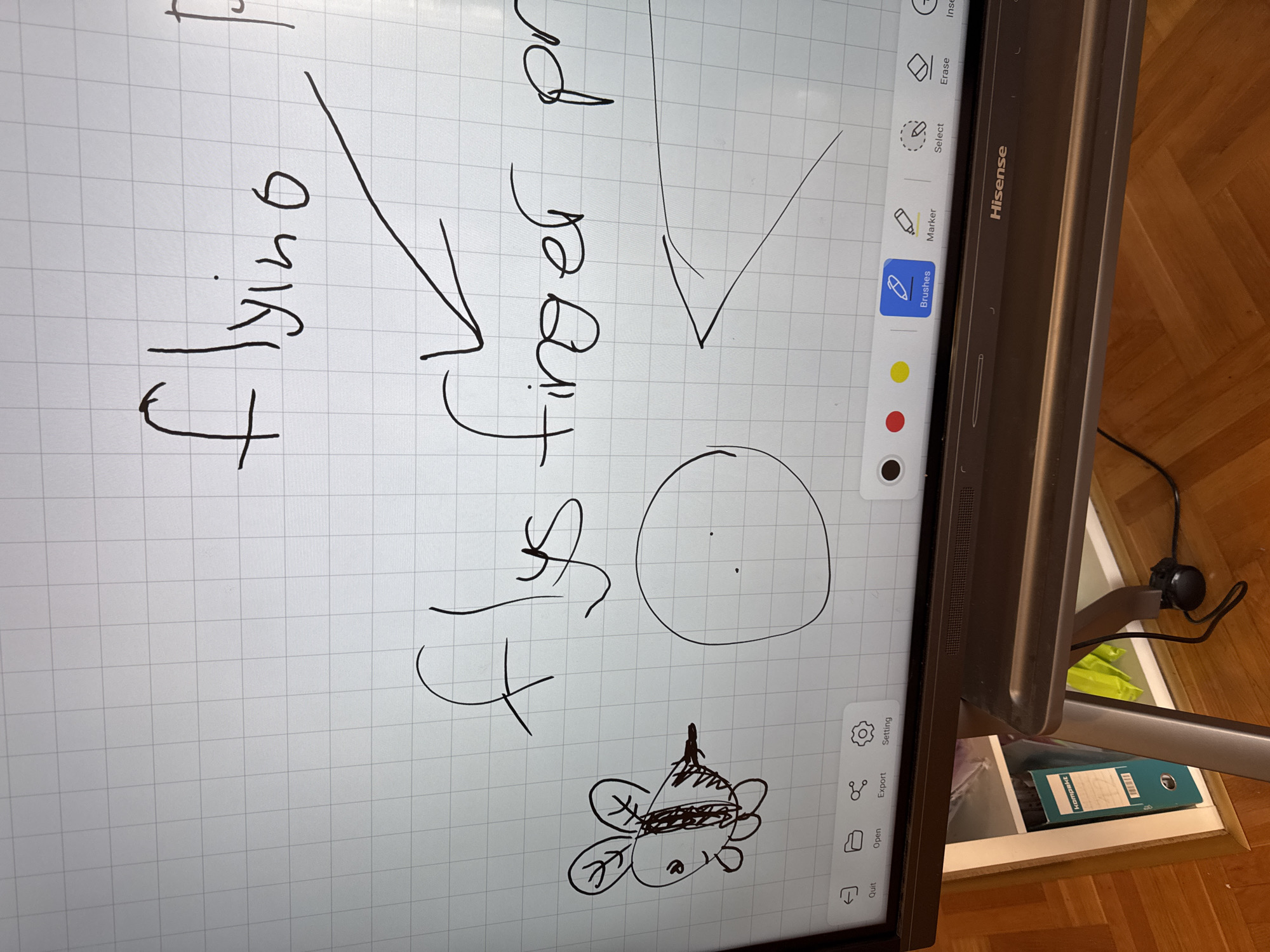 The image shows a close-up of a digital whiteboard with handwritten notes and drawings on it. The text reads "flying finger pro." There's a crude drawing of a bee below the text. The whiteboard is a Hisense brand and is mounted on a stand near a wooden floor. The whiteboard display shows an application with basic drawing tools, including a brush, marker, eraser, and select tool. There are also options to open, export, and adjust settings. A document can be seen in the background. The location in the city of Budva, Montenegro, cannot be determined from the provided image.