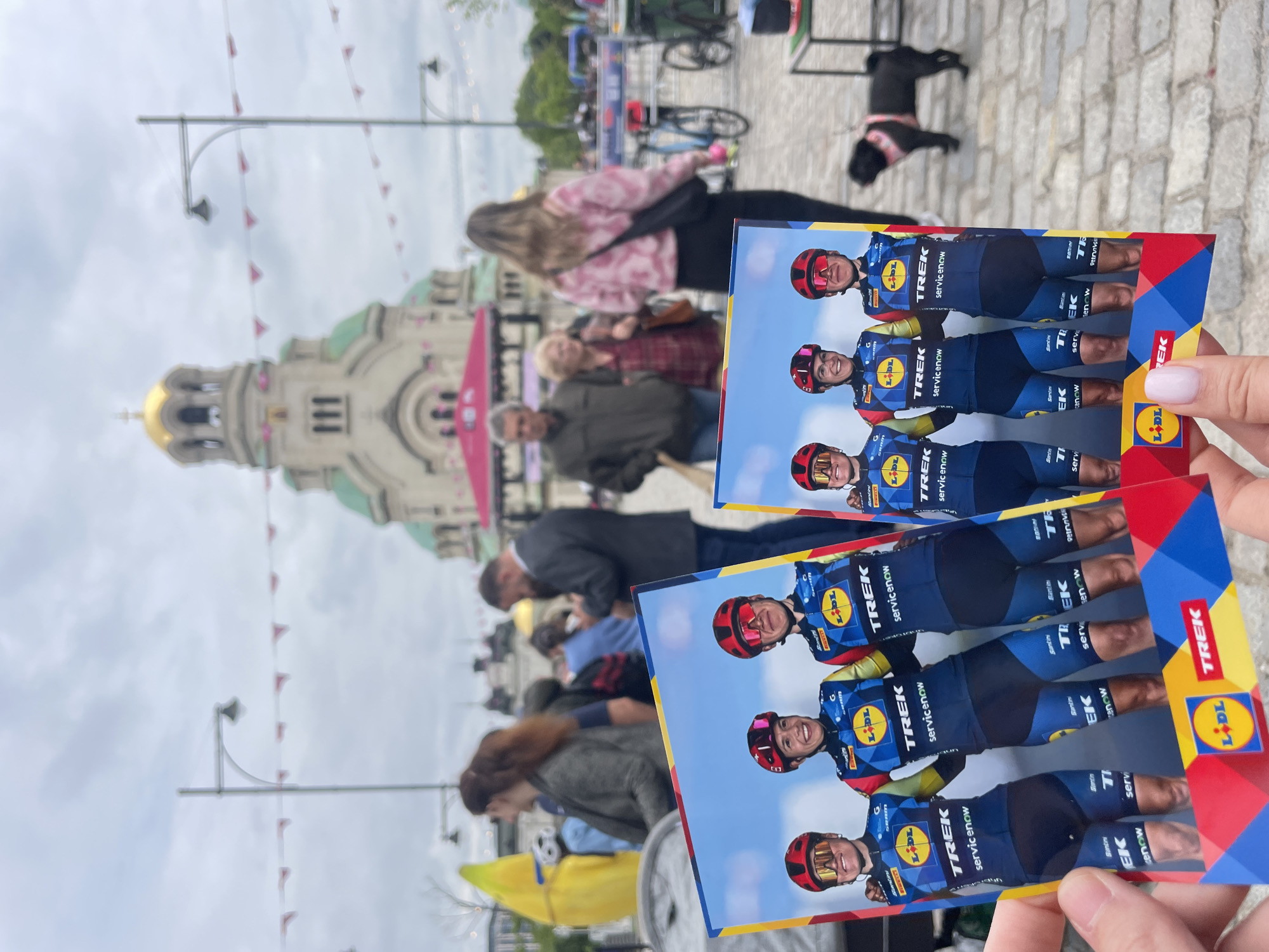 The photograph, taken at ground level, presents an outdoor urban scene in Sofia, Bulgaria. In the background stands the gold-domed Alexander Nevsky Cathedral. The sky is overcast.
The foreground features a pair of rectangular cards held in someone's hand. The cards display images of cyclists wearing blue and yellow uniforms, with the logos of Trek and Lidl prominently featured.
A diverse crowd of people populates the midground, suggesting a public event or gathering. A black dog is visible in the background, standing on a cobblestone sidewalk. Streetlights are positioned throughout the scene, adorned with small pink flags.