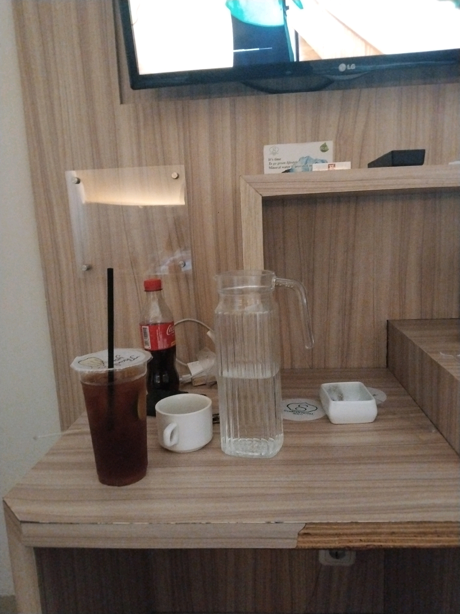 The image depicts a hotel room in Colomadu, Indonesia, specifically on a wooden table. On the table are various items including a plastic cup with a dark beverage and a straw, a white ceramic mug, a bottle of Coca-Cola, and a glass pitcher filled with water. A small white container is also visible. In the background, a television screen is mounted on a wooden wall panel. The overall scene suggests a moment of relaxation or refreshment in the hotel room.