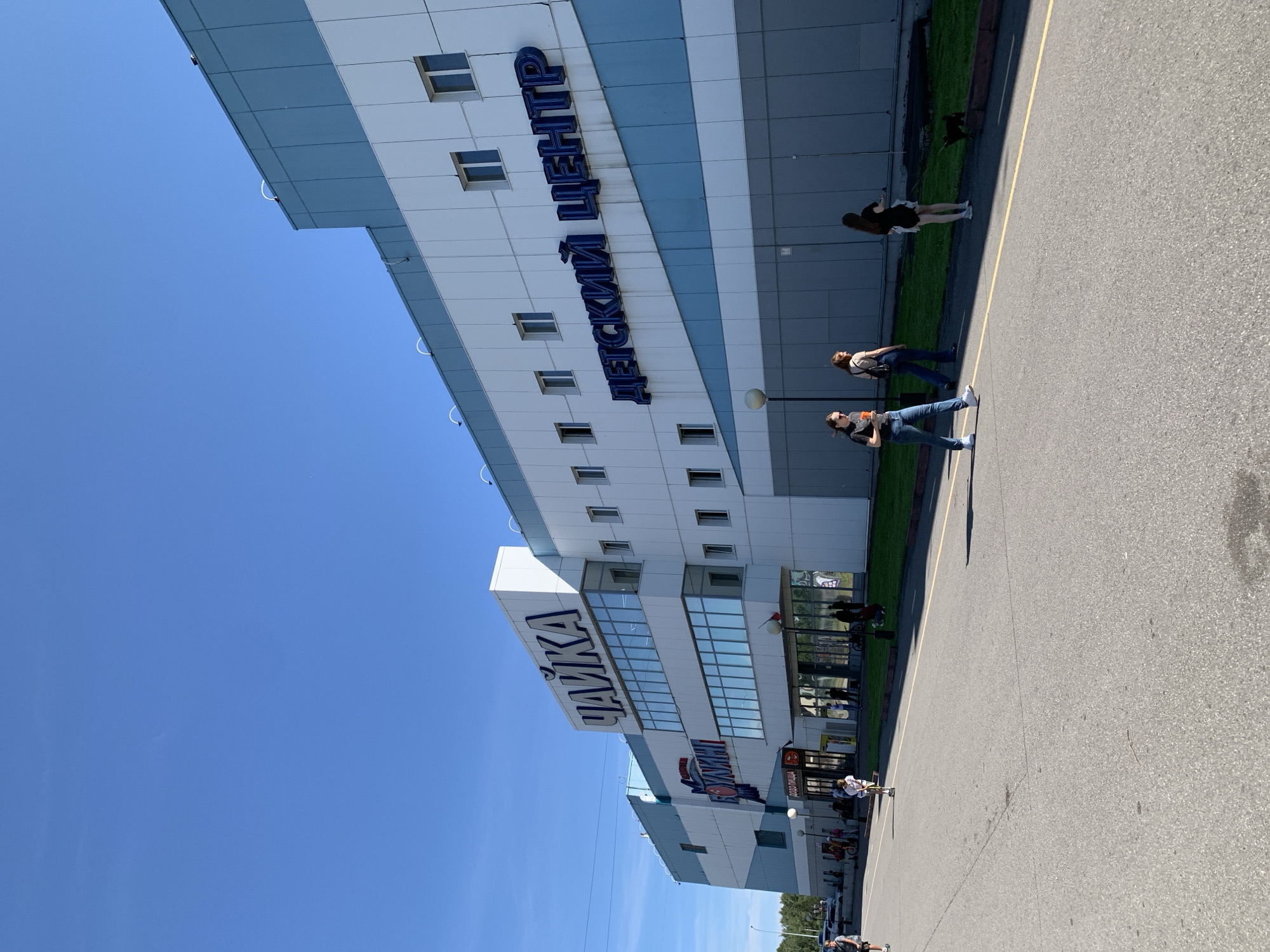 This is a full-shot, eye-level, outdoor, daytime, medium-angle photograph of a modern, light-colored building with blue accents, under a clear blue sky. The building appears to be a shopping center or a large public facility, with large windows and prominent signage. On the upper portion of the building, in large blue letters, is the Russian word "ЧАЙКА" (Chaika). Further to the right, also in large blue letters, is the Russian phrase "ДЕТСКИЙ ЦЕНТР" (Detskiy Tsentr), meaning "Children's Center". In the foreground, a paved area, likely a sidewalk or parking lot, stretches across the bottom of the frame. Several people are visible: three women are walking on the paved area from left to right, one of whom is walking a small black dog on a leash. In the middle ground, several individuals are near the entrance of the building, some appearing to be entering or exiting. The building itself has a contemporary architectural style, with multiple stories and sections. The environment appears to be urban, with a hint of greenery and trees visible in the background to the left. The overall impression is one of a sunny, active public space. The image is located in Prokopyevsk, Russia, as identified by the signage on the building.