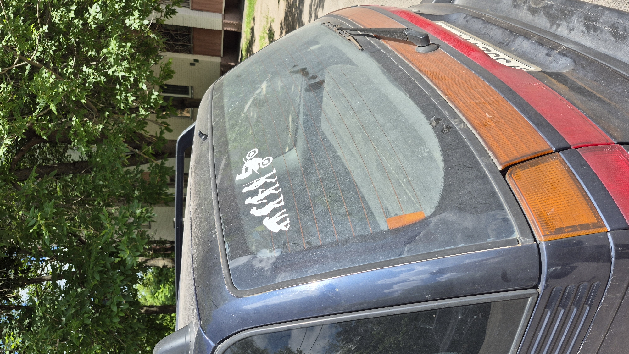 A dark blue station wagon is parked on a paved surface in front of a building with a light-colored facade and trees with green foliage. The car's rear window has a white decal depicting the evolutionary stages of man, culminating in a figure riding a motorcycle. The car appears dusty. The license plate is partially visible. The scene is lit by daylight. The location is Homyel, Belarus.