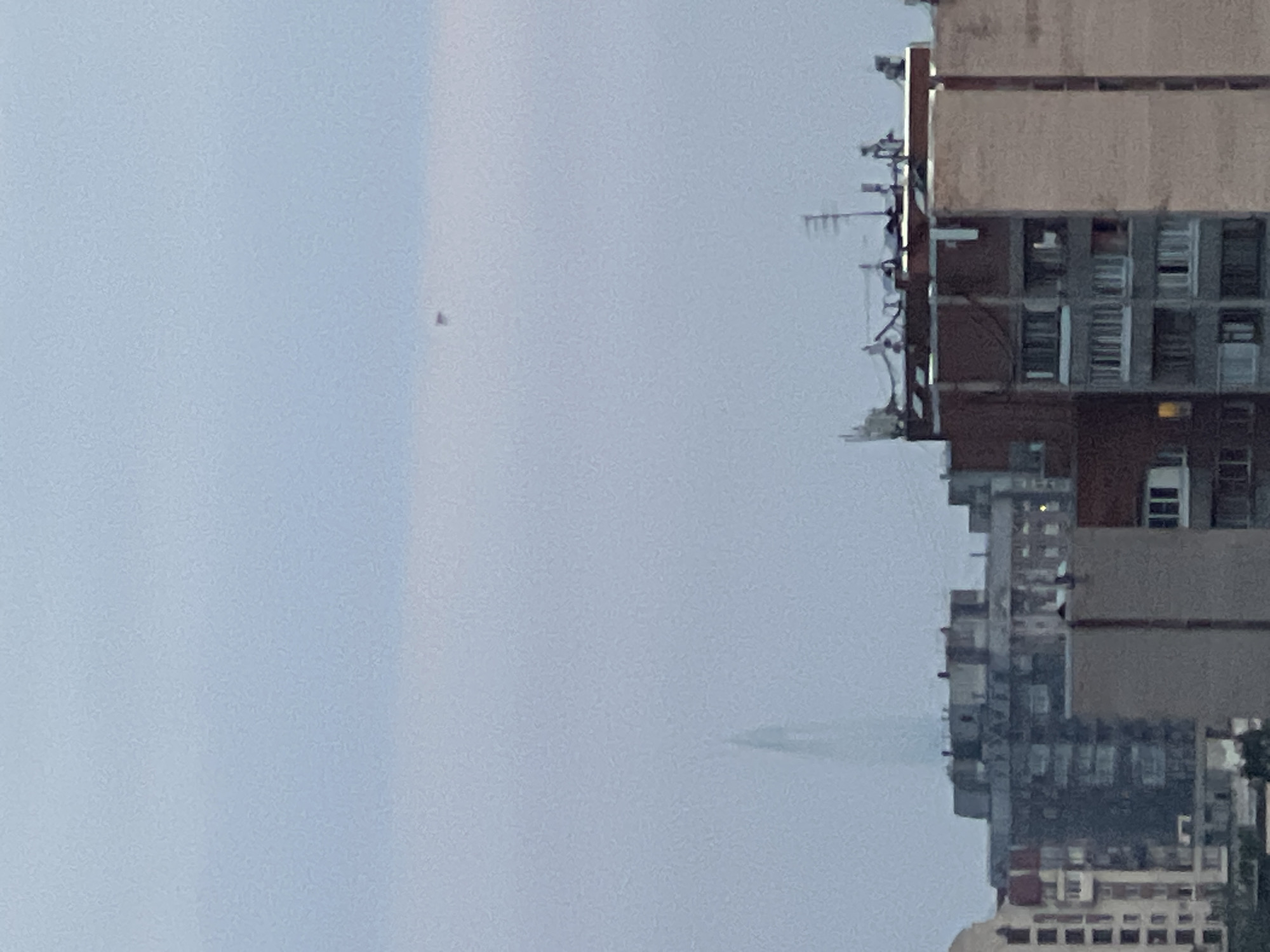The image captures an urban skyline under a hazy, overcast sky. In the foreground, various apartment buildings are visible, exhibiting different architectural styles and ages. Some buildings have multiple stories with visible windows, while others are more weathered. Antennas and other rooftop structures are also present on some of the buildings. In the mid-ground, a distinct, tall, spire-like building with a translucent, possibly glass, facade can be seen rising above the lower structures. In the upper portion of the frame, a small, dark, indistinct object is suspended in the sky, appearing as a speck against the light blue haze. The overall impression is of a dense urban environment, likely a city, with a prominent modern skyscraper in the distance and a curious aerial anomaly.