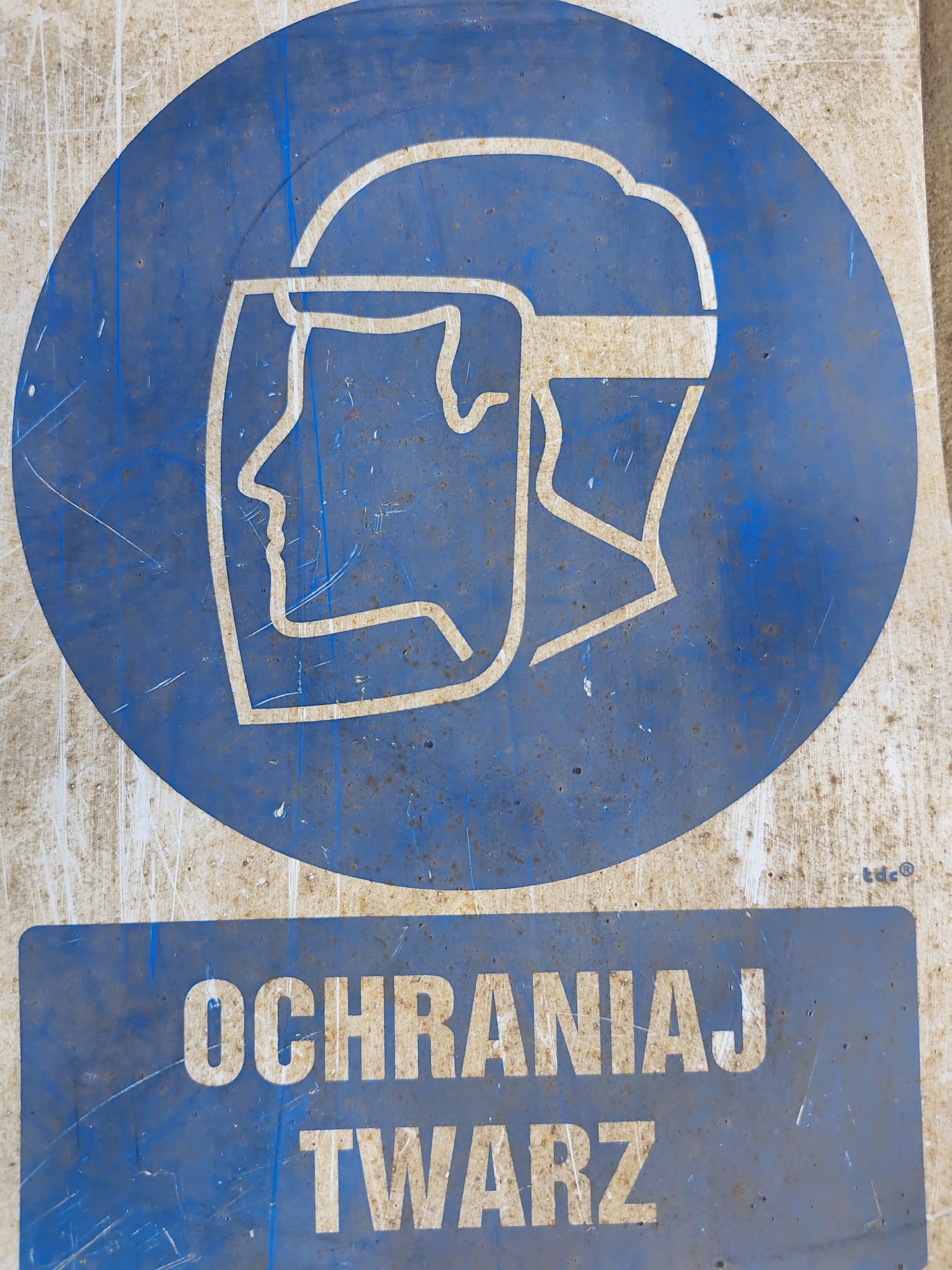 This is a close-up, eye-level, vertical shot of a weathered blue and white safety sign. The sign is located outdoors, as evidenced by the light, and appears to be mounted on a rough, textured surface. The primary graphic on the sign is a white outline of a person wearing a face shield, indicating the need for facial protection. Below this graphic, also in white lettering on a blue background, are the Polish words "OCHRONIAJ TWARZ," which translates to "Protect Face." The sign shows signs of age and wear, with rust spots, scratches, and peeling paint visible on its surface. The text "tdc®" is faintly visible in the lower right corner of the blue circular section. The context of "Police, Poland" is provided externally and is not discernible from the image itself, but suggests this sign is a public safety notice within that city.