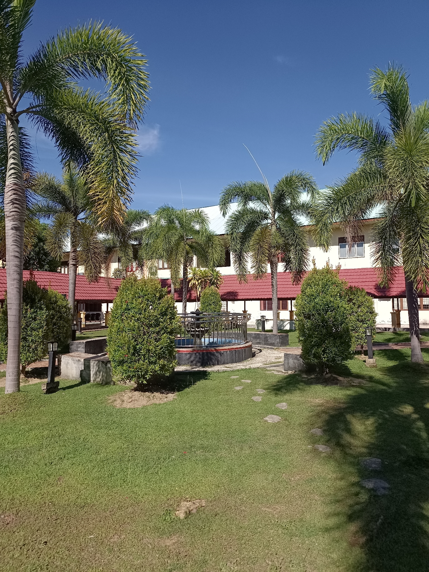 The image shows a tranquil courtyard area with lush green grass, surrounded by palm trees and manicured bushes. In the background, a multi-story building with a red tiled roof and cream-colored walls stands under a clear blue sky. A small, decorative fountain is visible in the center of the courtyard, enclosed by a black metal fence. Several stone slabs are laid out on the grass, forming a pathway. The scene is well-lit, suggesting it's a sunny day in Mesjid Reuntoh, Indonesia. There are no people present in the image.