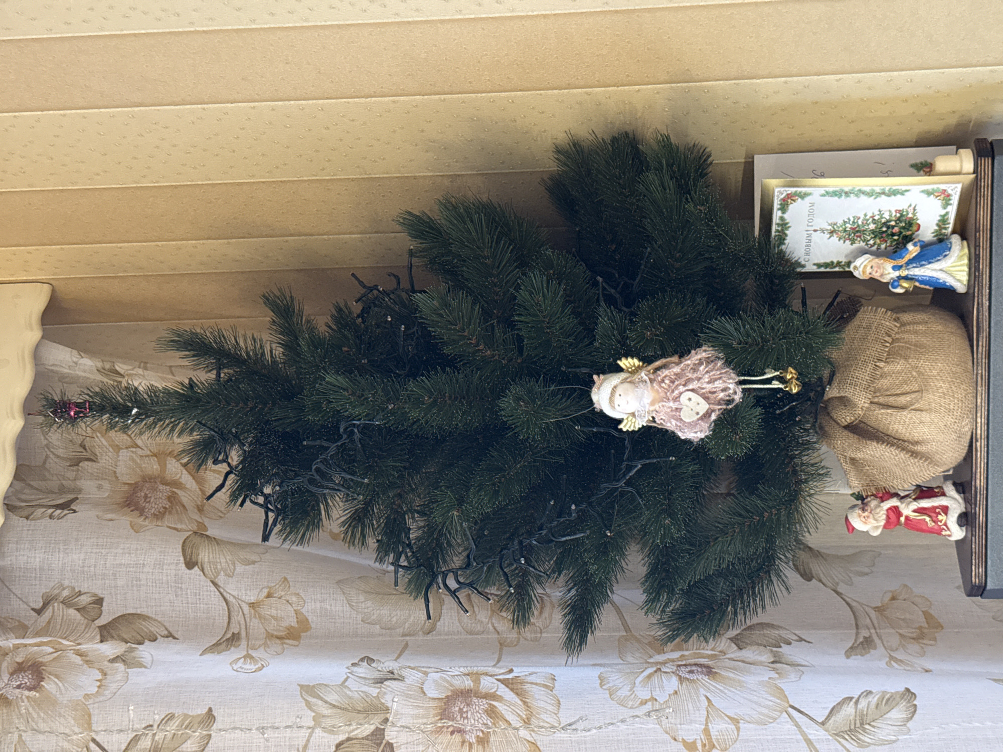The image shows a close-up view of a partially decorated artificial Christmas tree lying on its side. The tree has dark green pine needles and is adorned with a string of white fairy lights. A small, light pink knitted angel with gold wings is attached to one of the branches, and further down, a small red sack, possibly a gift bag, is visible. To the right of the sack, a miniature Santa Claus figurine in a red and white suit is placed. Behind the tree, there is a dark wooden box or shelf. Inside the box, a Christmas card with a depiction of a decorated Christmas tree and text in Russian, "С НОВЫМ ГОДОМ" (Happy New Year), is propped up. A small figurine of a woman in a blue coat and hat, possibly Snegurochka (the Snow Maiden), stands next to the card. The background above the tree appears to be a textured, cream-colored ceiling with horizontal paneling. The lower portion of the image shows a fabric with a floral pattern in shades of beige and cream. The lighting suggests an indoor setting.