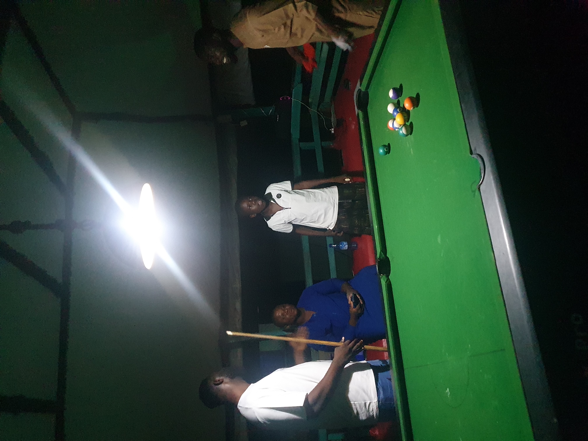The image depicts an indoor billiard hall, possibly at a recreational facility or a private residence, located in Lokoja, Nigeria. Four individuals are present. In the foreground, a green billiard table is illuminated by an overhead light fixture. Several billiard balls are scattered on the table. To the left, a man in a white t-shirt and blue shorts stands holding a billiard cue. To his right, a person in a blue garment is seated, looking at the table. Further to the right, a young man in a white polo shirt and patterned shorts stands, and to his right, another man in a light brown outfit is visible. The environment appears to be a covered outdoor space or a room with dark walls and ceiling, with a railing in the background. The lighting is dim, except for the direct illumination from the overhead lamp.
