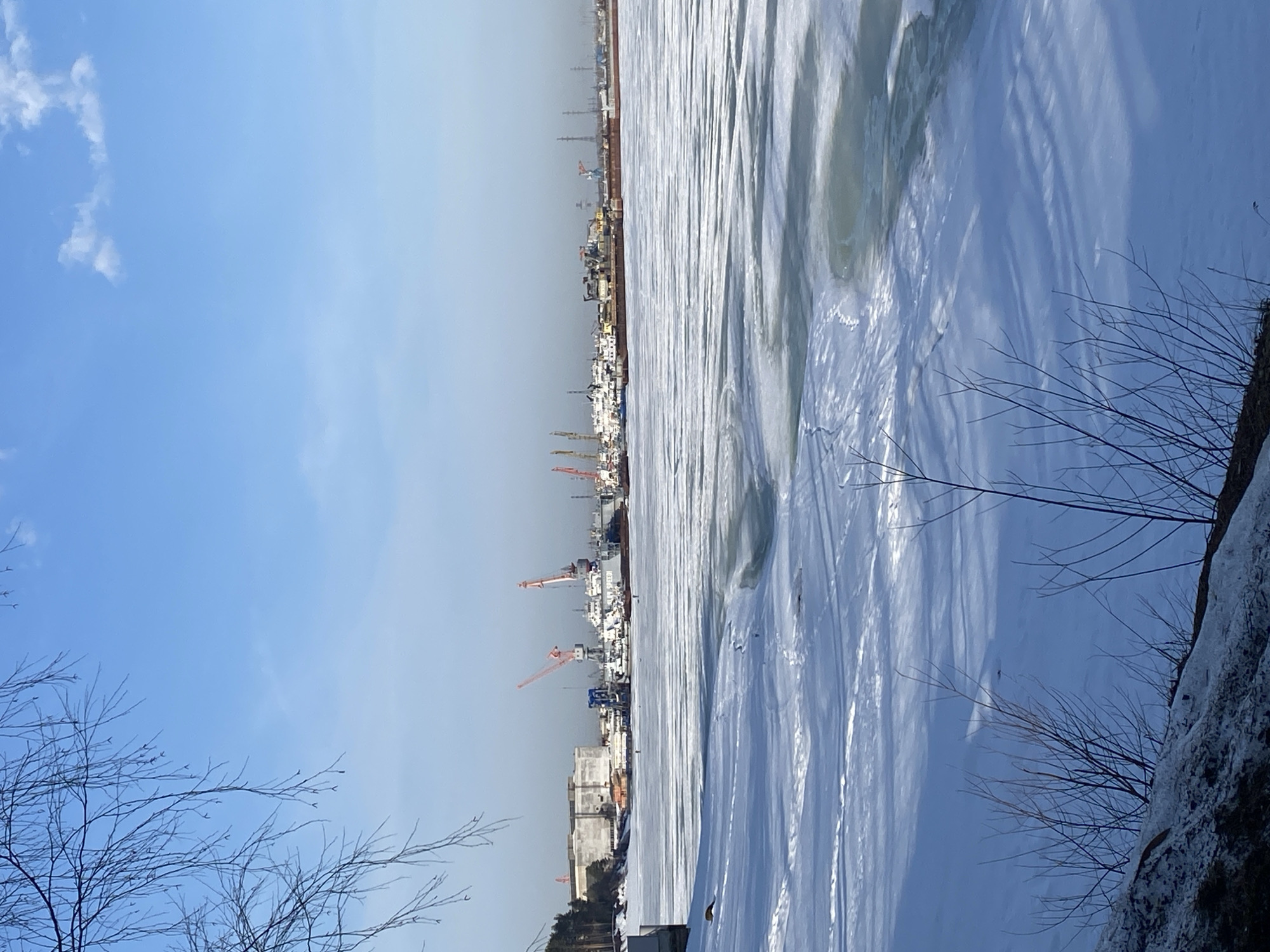 The image depicts a partially frozen body of water, possibly the Kama River, in Chaikovsky, Russia, bordered by industrial facilities and docks. The water's surface is covered with a layer of ice, showing a cracked and uneven texture. In the background, cranes, buildings, and structures characteristic of a port or industrial area are visible. The sky is light blue with some wisps of clouds. Bare tree branches are present in the foreground.