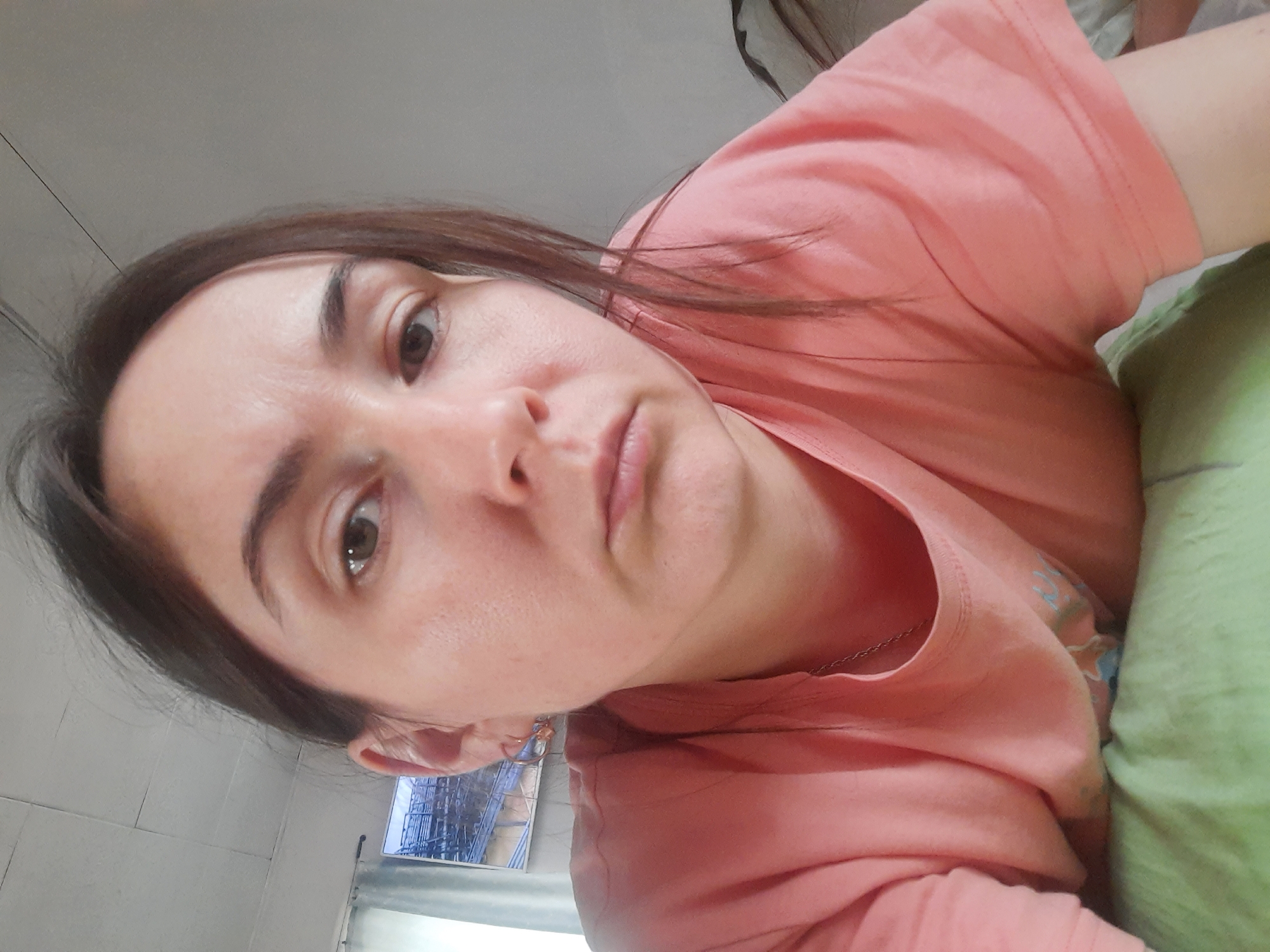 A person is visible in this close-up, head-and-shoulder shot. The individual has fair skin, brown hair, and brown eyes. They are wearing a light pink t-shirt and appear to be resting on a green pillow. Part of a white tiled wall and a window with a view are in the background. Based on the location provided, this scene is purportedly in Krasnogorskoye, Russia. However, no details in the image definitively confirm the specific location.
