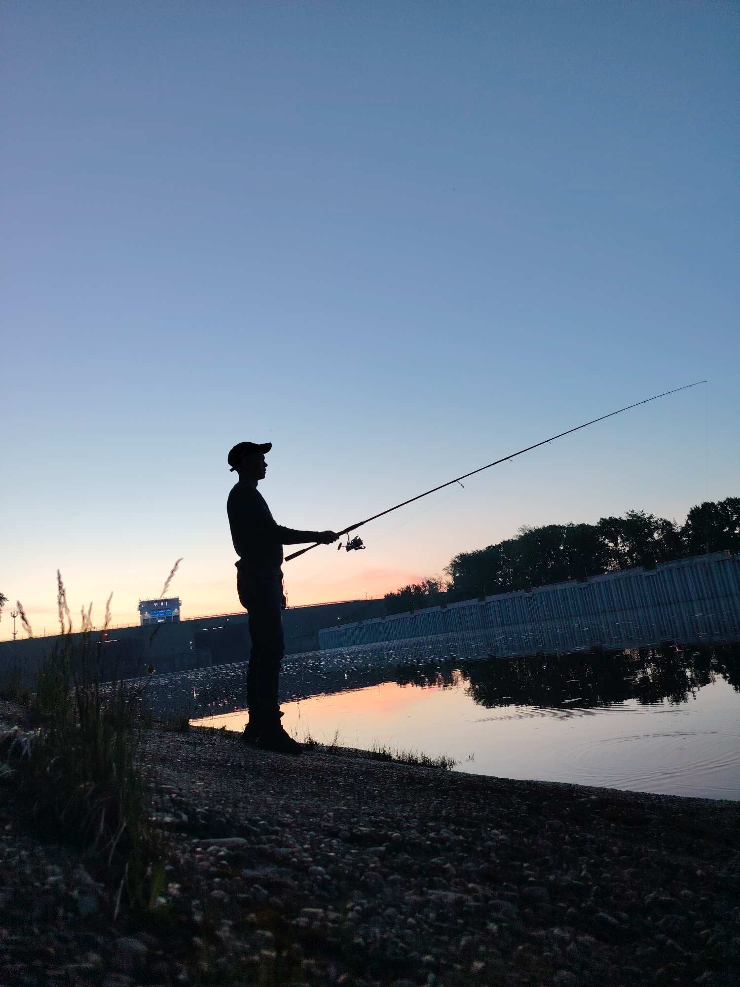 The image shows a silhouette of a person fishing at dusk on the bank of a body of water. The individual is standing, wearing a cap, and holding a fishing rod, which extends towards the right side of the frame. The background includes a concrete structure, possibly a dam or reinforcement, and trees along the water's edge. The sky transitions from a light blue at the top to a warm orange near the horizon, reflected in the water. The shoreline is covered in pebbles and vegetation. The location is likely in Chaikovsky, Russia.