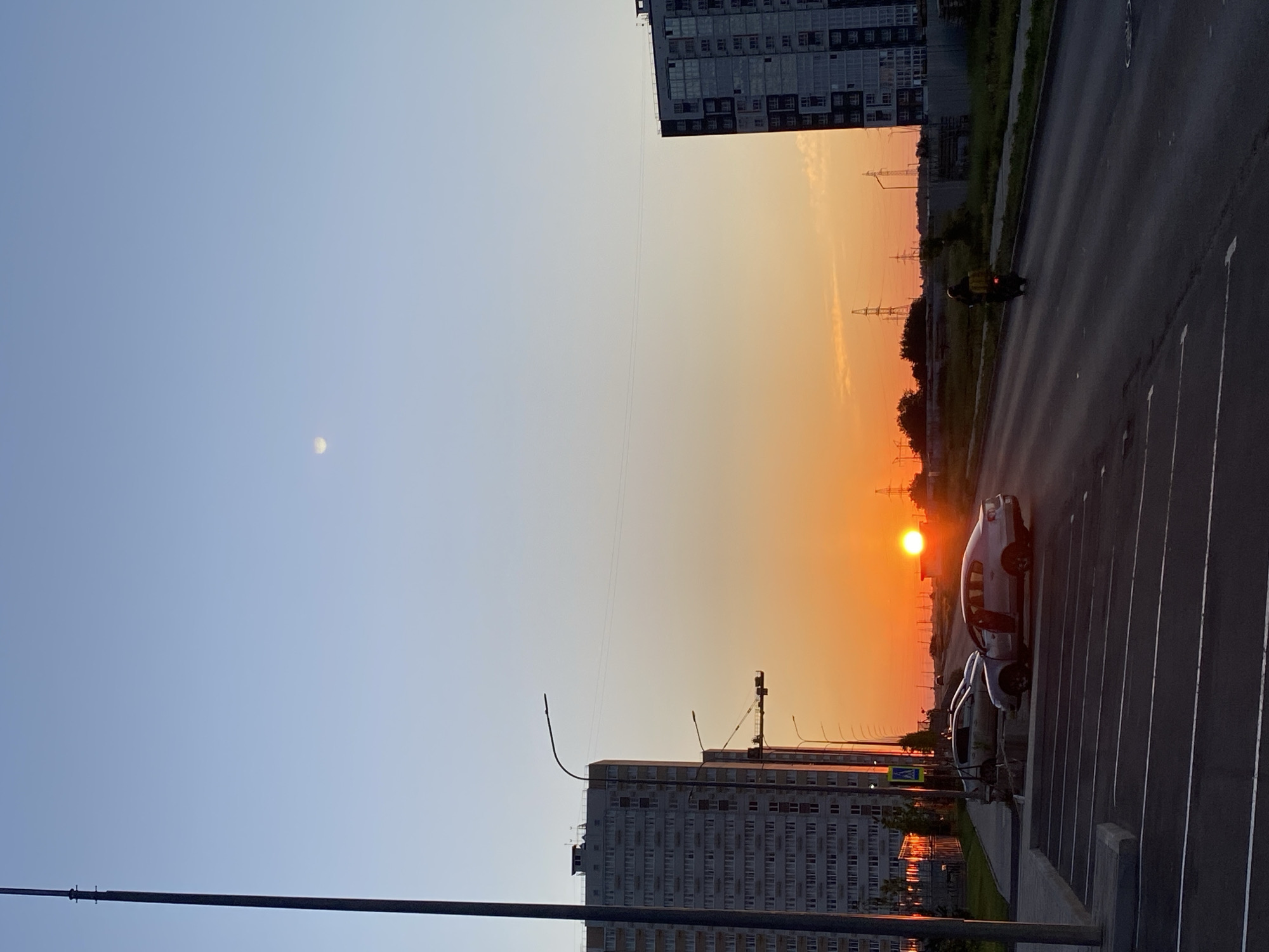 An outdoor, full shot captures a cityscape at sunset. The sky is a gradient of soft blue at the top, transitioning to a vibrant orange near the horizon. The sun is a bright, white orb positioned above a road. Two tall apartment buildings flank the scene, one on the left and one on the right, with multiple windows visible. A dark pole stands on the far left, extending vertically from the bottom to the top of the frame. Several streetlights are scattered throughout the image, and power lines are visible in the distance. A road runs diagonally across the middle of the frame, with a parked silver car and a motorcycle with a yellow box on the back on it. The ground in the foreground is an asphalt parking lot with white parking lines. A faint moon is visible in the upper right quadrant of the sky.