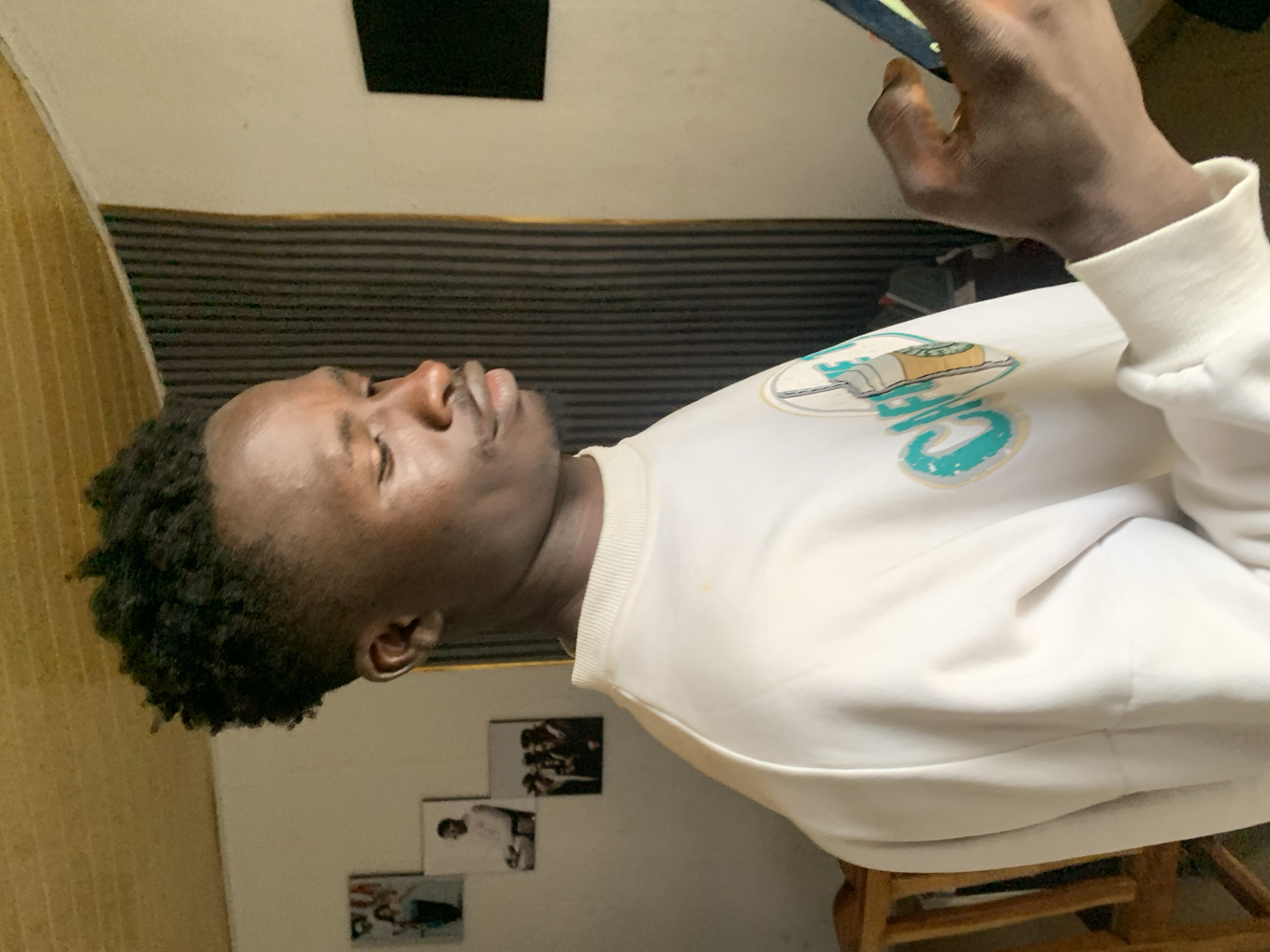 A Black male is shown from the shoulders up, in a profile view, looking down at a smartphone he is holding in his right hand. He has dark, curly hair styled in a high-top fade and is wearing a white sweatshirt with a graphic on the left chest that appears to be a stylized coffee cup with the word "CAFE" below it. The man's eyes are closed or partially closed. He is indoors, in what appears to be a residential setting. Behind him, on the wall, are several framed pictures. To his left, a wooden chair is visible. The background features a ribbed, dark-colored vertical surface and a plain wall. The lighting suggests it is daytime, with light coming from the right side of the frame. The overall impression is of a casual, personal moment captured indoors, possibly in Jos, Nigeria, based on the provided context.