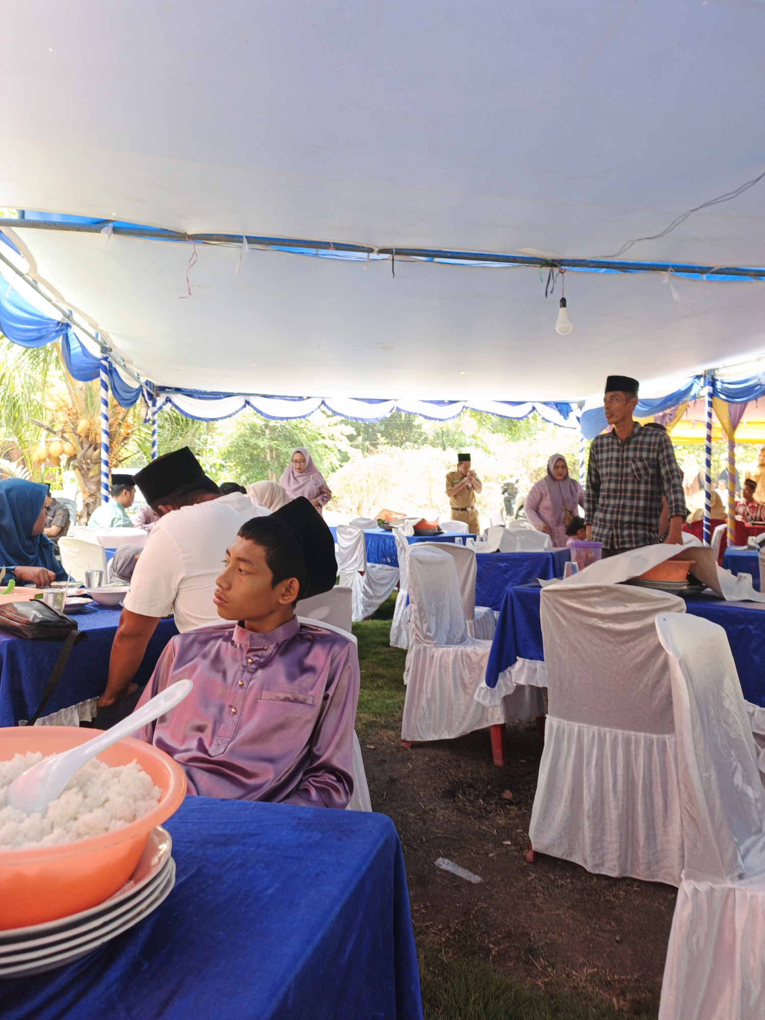 In Pematang Duku, Indonesia, an outdoor event is underway beneath a white tent with blue and white draped awnings. Tables covered in blue cloths are set with white chairs draped in white cloths. A young man wearing a purple satin shirt and a black peci (cap) is seated at a table in the foreground, looking to the right. A white bowl containing white rice and a white spoon is placed on stacked plates in front of him. In the mid-ground, a man wearing a black peci and a white shirt is visible from the back, facing away from the camera, with another person in a black peci next to him. Further in the background, several other individuals are present, including a woman in a pink hijab and a man in a plaid shirt. The ground is covered with dirt and some grass.