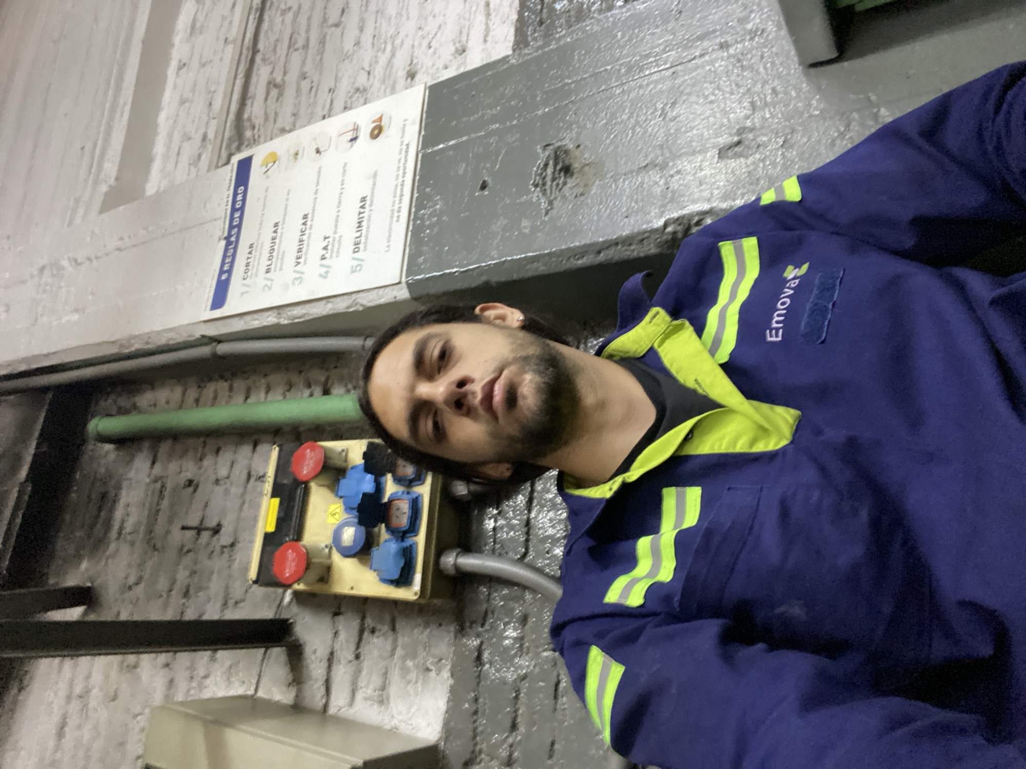 A male individual is captured in a close-up selfie within an indoor industrial environment. The subject has dark hair, a light beard, and wears a dark blue work uniform featuring neon green accents and reflective strips. The uniform prominently displays the "Emova" logo and text on the left chest. He directly faces the camera with a neutral expression. Behind the subject, a white-painted brick wall occupies the upper left, transitioning to a grey-painted wall or column on the right. Mounted on the brick wall to the subject's left is a yellow electrical distribution panel containing two red and at least three blue industrial power outlets, along with a yellow triangular electrical hazard symbol. A silver conduit extends downwards from this panel. To the subject's right, a white safety sign titled "5 REGLAS DE ORO" (5 Golden Rules) is affixed to the grey wall, listing five numbered safety steps in Spanish, including "CORTAR" (Cut) and "BLOQUEAR" (Lock out). The sign also displays a warning message about the dangers of electricity. A vertical green pipe is visible on the far right of the frame, and a black metal beam is present in the upper left. The "Emova" branding identifies the location as a facility associated with the Buenos Aires Subte (subway) system, likely a maintenance depot or operational area within Buenos Aires, Argentina.