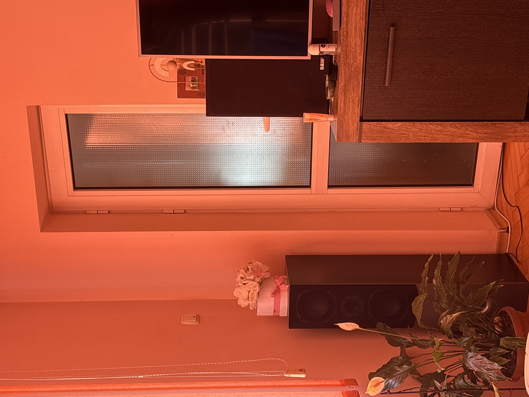 The image shows an interior view with a window in the center. Above the window is a television set and a cabinet. Below the window, there is a speaker and a potted plant. Next to the speaker, there's a gift with a pink ribbon and a flower bouquet. The wall color is orange. The location is Panagyurishte, Bulgaria.