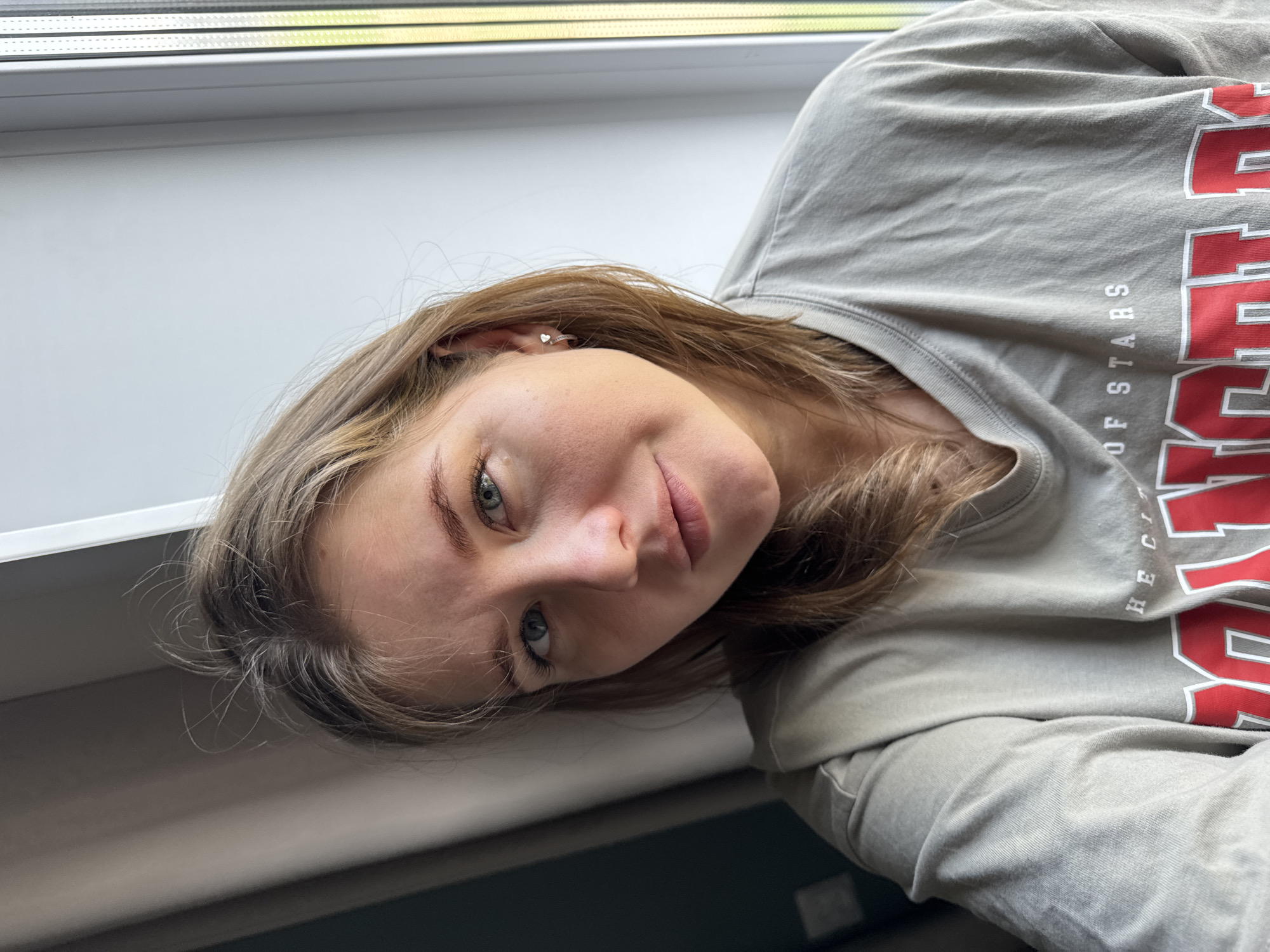A young woman with fair skin, light brown hair, and blue-grey eyes poses in front of a window with a white frame. She is wearing a light gray, long-sleeved shirt with the word "RIDER" in large, red, capitalized letters and "THE CITY OF STARS" in smaller, white capitalized letters. She is wearing a small, heart-shaped earring in her left ear. The location is possibly Ферма, Russia, but this is not confirmed by the image.