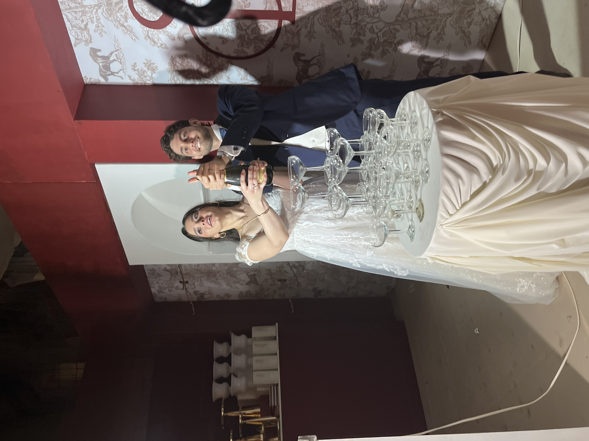 A wedding scene is captured indoors in what appears to be Orizaba, Mexico, judging from possible architectural and decorative styles. A couple, dressed in wedding attire, is positioned in front of a tower of clear wine glasses placed on a round, white table. The man, wearing a dark suit, is holding a champagne bottle and assisting a woman in a white wedding dress to open the bottle, presumably to pour into the glasses. The backdrop includes a textured wallpaper or wall design in muted tones. The location appears to be a reception venue with dark walls and architectural details.
