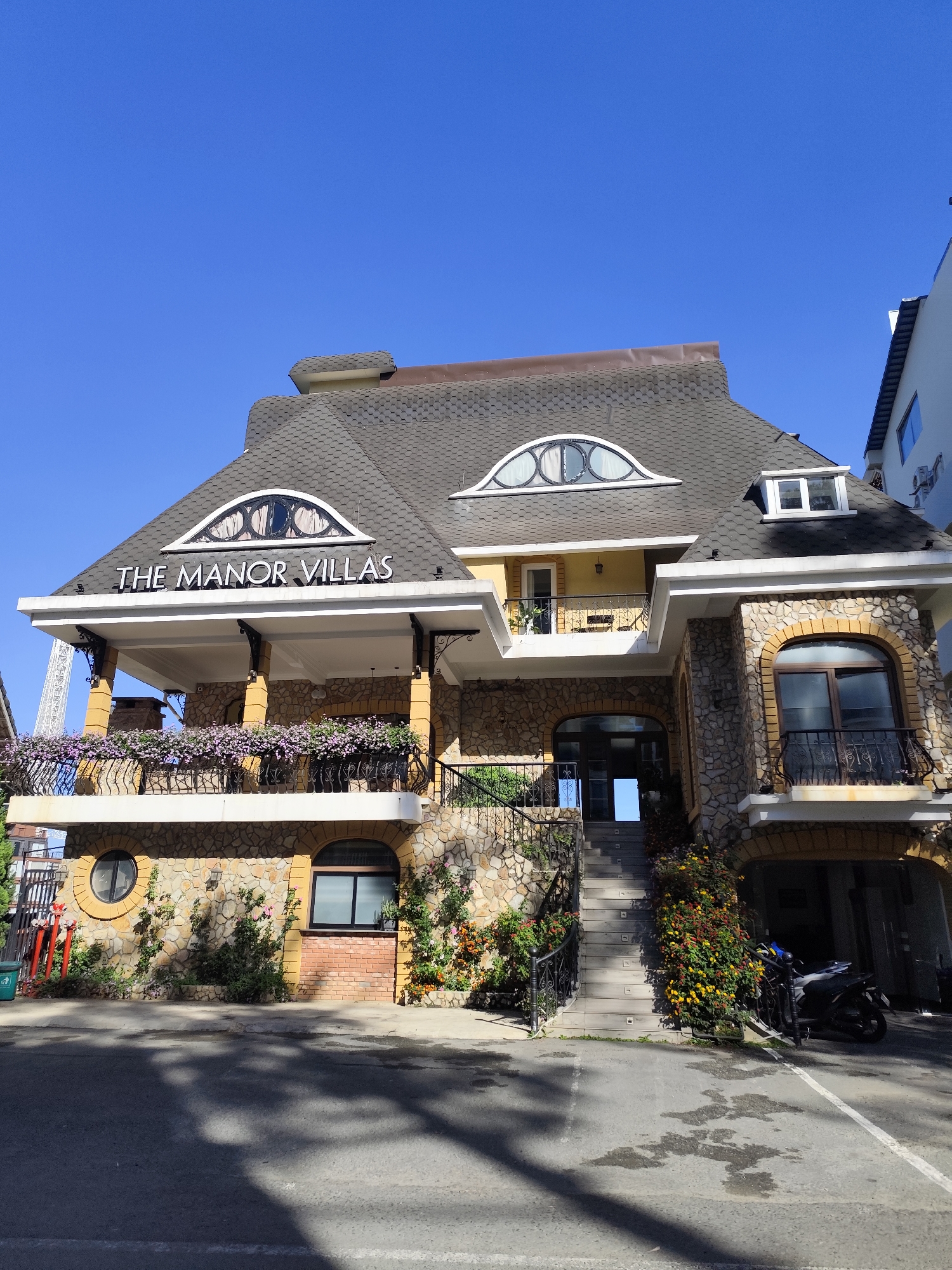 The image depicts the exterior of "The Manor Villas" in Da Lat, Vietnam, under a clear blue sky. The building features a distinctive architectural style with a steeply pitched, gray shingled roof and a facade incorporating stone and brick elements. The name "The Manor Villas" is prominently displayed on a white sign above a covered balcony adorned with flowering plants. Architectural details include arched windows and doors, wrought iron railings, and decorative columns.
A set of stairs leads up to the main entrance, with vegetation growing along the sides. A motorcycle is parked beneath an arched section of the building. The foreground shows a paved surface, likely a street or parking area, with shadows indicating the time of day. No people are visible in the image.