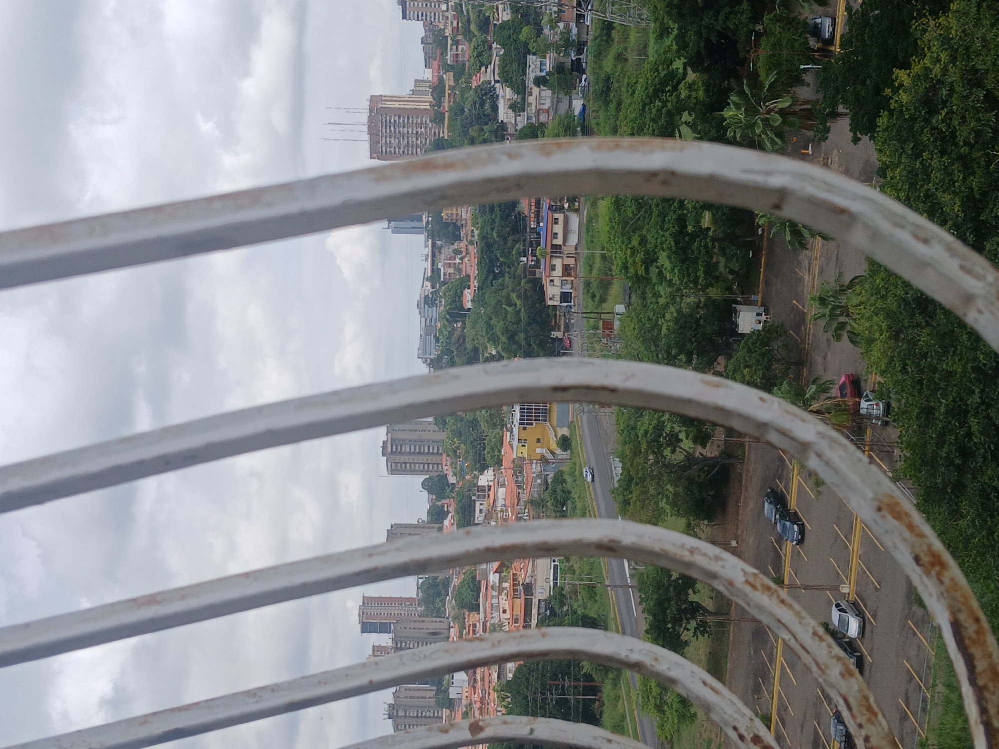 This image provides a medium-distance view of a cityscape in Puerto Ordaz, Venezuela, partially obscured by the foreground of weathered, white metal bars. The scene depicts a dense urban environment with a mix of residential and commercial buildings, characterized by red-tiled roofs common in the region. Several multi-story apartment buildings and a few taller structures are visible in the background. Green trees and vegetation are interspersed throughout the city, indicating a well-treed urban landscape.
In the middle ground, a road with visible lane markings runs through the city, and a single white car is observed traveling on it. A parking lot is situated in the lower portion of the image, with several cars parked in designated spots. These vehicles appear to be typical passenger cars. The sky is overcast with gray clouds, suggesting a cloudy or possibly rainy day. The overall impression is a typical day in a Venezuelan city.
