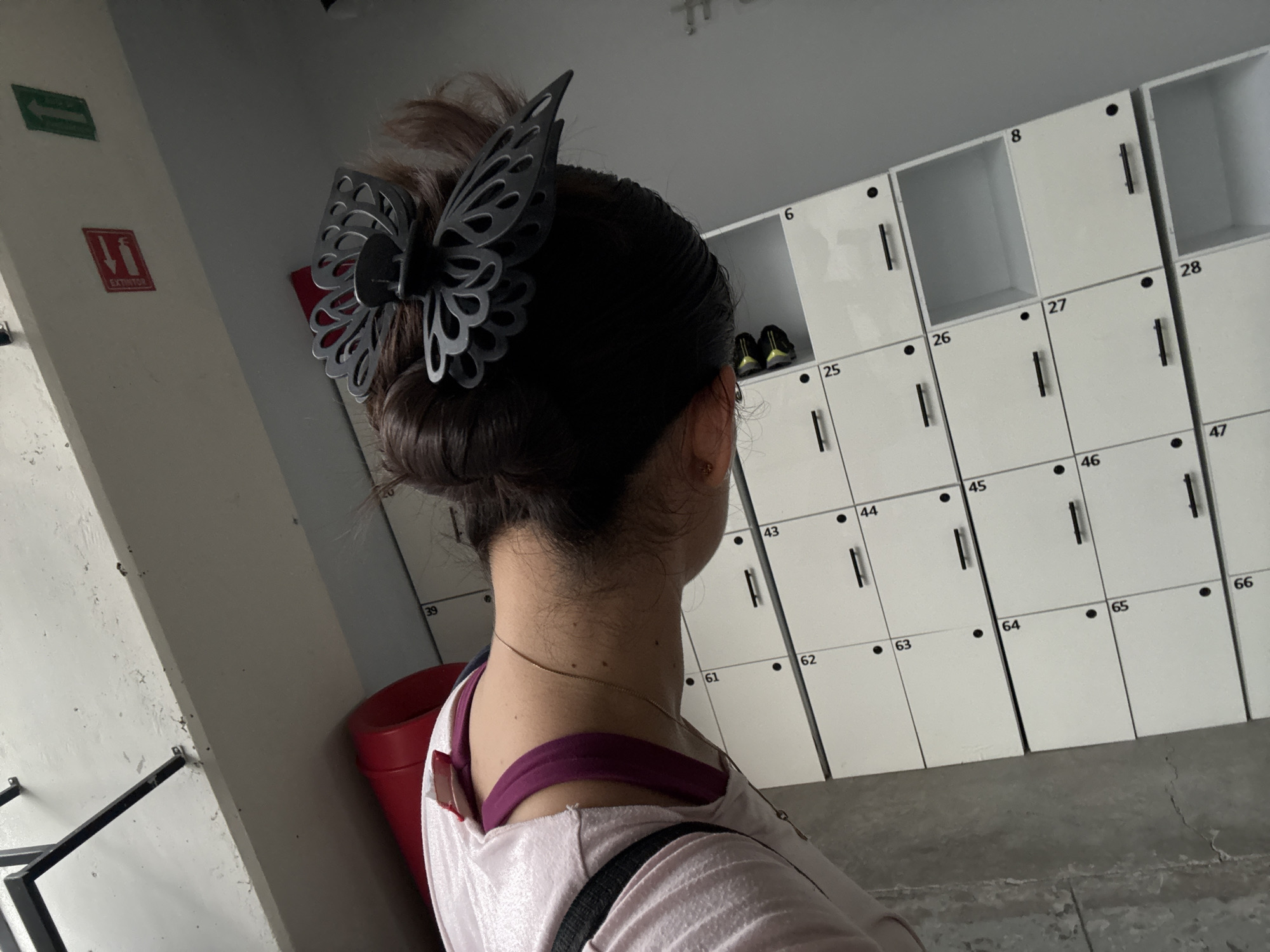 A woman with dark hair styled in a bun with a black butterfly clip is facing away from the camera. She is wearing a light pink top with a visible purple strap and a thin gold necklace. A black bag strap is visible across her shoulder.
Behind her are white lockers with black handles and numbers ranging from 6 to 66. A red fire extinguisher container is visible on the left, near a wall with a green arrow sign and a red fire extinguisher sign. The scene appears to be a gym or changing room, possibly located in Puebla, Mexico, as inferred from the EXINTOR sign. A metal rack is visible on the lower left. The floor is made of concrete.