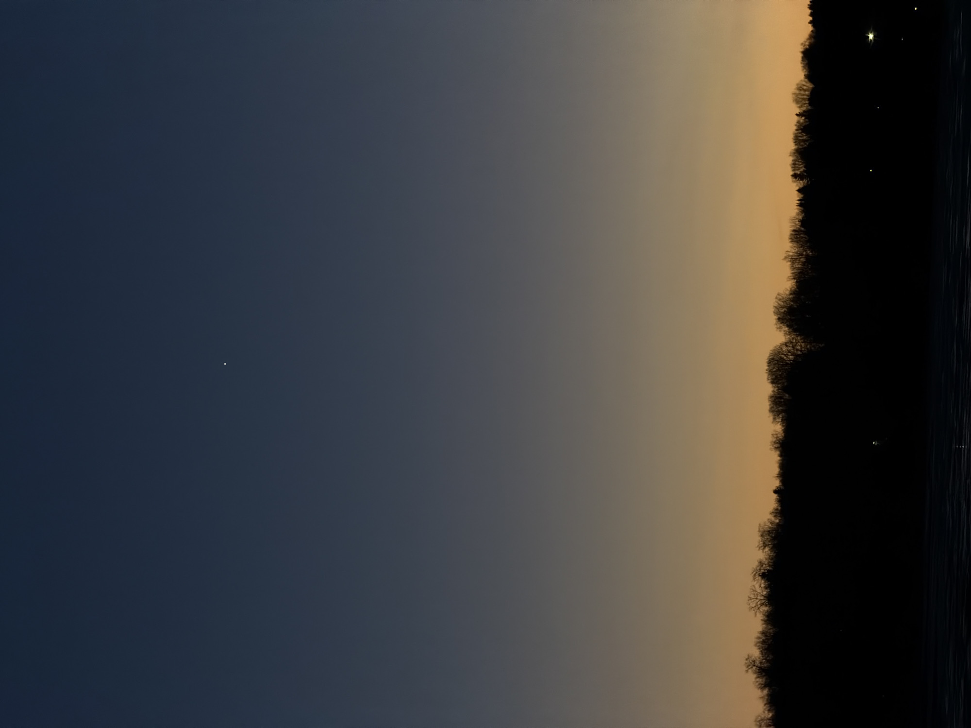 The image shows a dusk scene in Izhevsk, Russia. The sky transitions from a dark blue on the left to a pale orange near the horizon on the right. A single bright celestial object is visible in the dark blue portion of the sky. A dark silhouette of trees runs along the right edge of the image, with several lights visible through the trees and in the water, suggesting a waterfront location. The photograph is rotated 90 degrees clockwise.