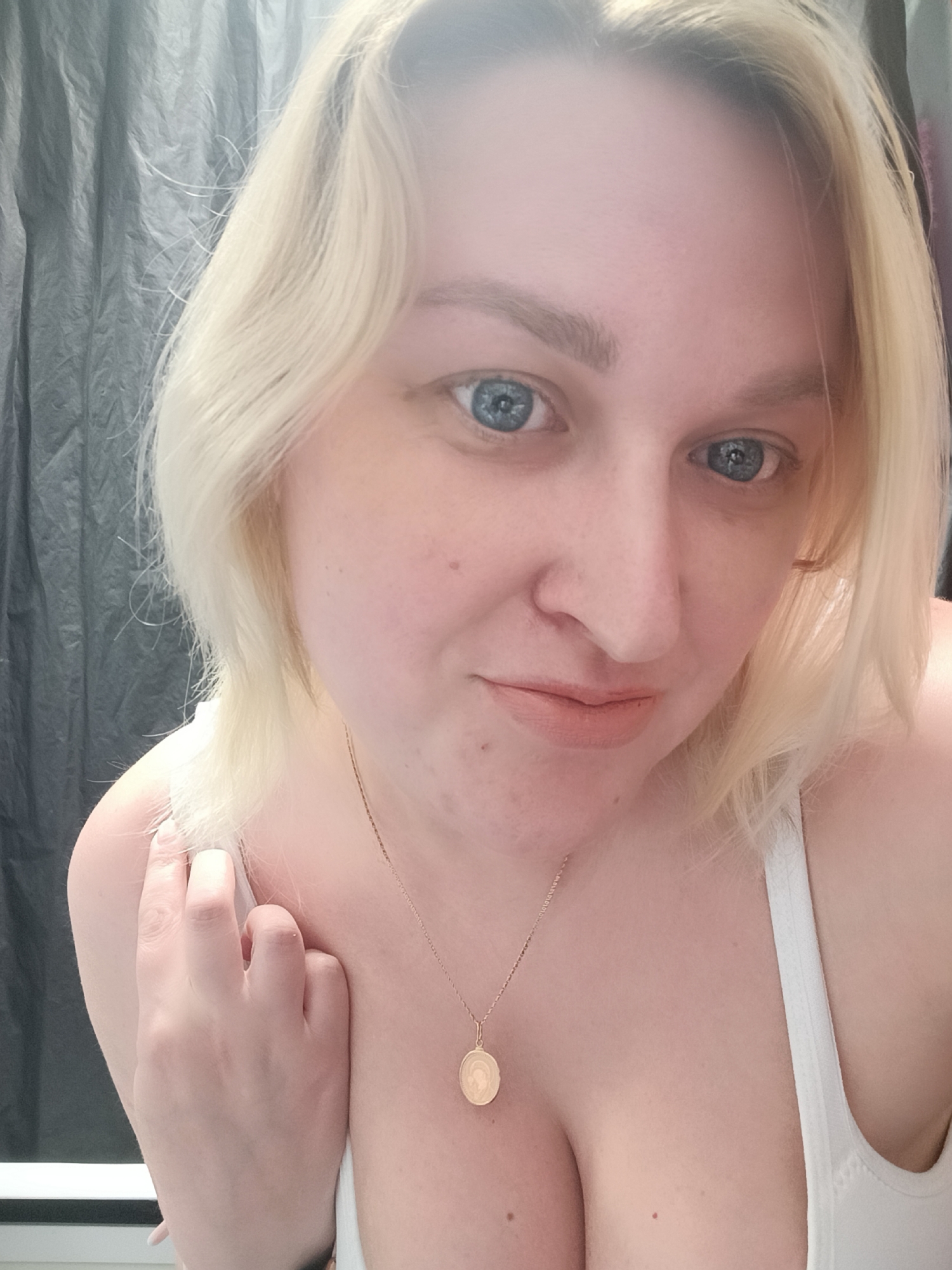 A close-up portrait features an adult individual with fair skin, bright blue eyes, and light blonde, shoulder-length hair. She looks directly at the camera with a slight, closed-mouth smile, conveying a calm demeanor. Her right hand is gently placed on her left shoulder, near her hair. She is wearing a white top, possibly a tank top or bra, and a delicate gold-colored chain necklace with a circular pendant. The background is a dark, textured material, likely a gray or black fabric curtain. Soft, diffused lighting illuminates the scene, suggesting an indoor setting during daytime. A faint light-colored horizontal line is visible in the lower left background, potentially a window sill or another fixture in the room. The image captures a casual moment. No visible text is present. The context provided indicates the location is Chaikovsky, Russia.