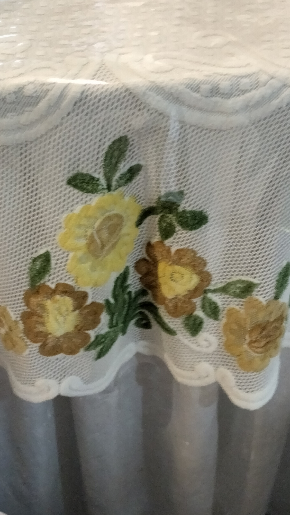 The image shows a close-up of a white lace tablecloth with embroidered flowers. The tablecloth has a scalloped edge with intricate patterns. The embroidery features several flowers in shades of yellow, brown, and green leaves. The flowers appear to be a mix of rose and daisy-like shapes. The background is blurred, but suggests a surface, possibly a table, covered by the tablecloth, and a gray fabric underneath, possibly a curtain or the tablecloth's lining. The lighting is soft and even, indicating an indoor setting, possibly during the day. There is no visible text in the image. The overall impression is one of delicate craftsmanship and home decor.
