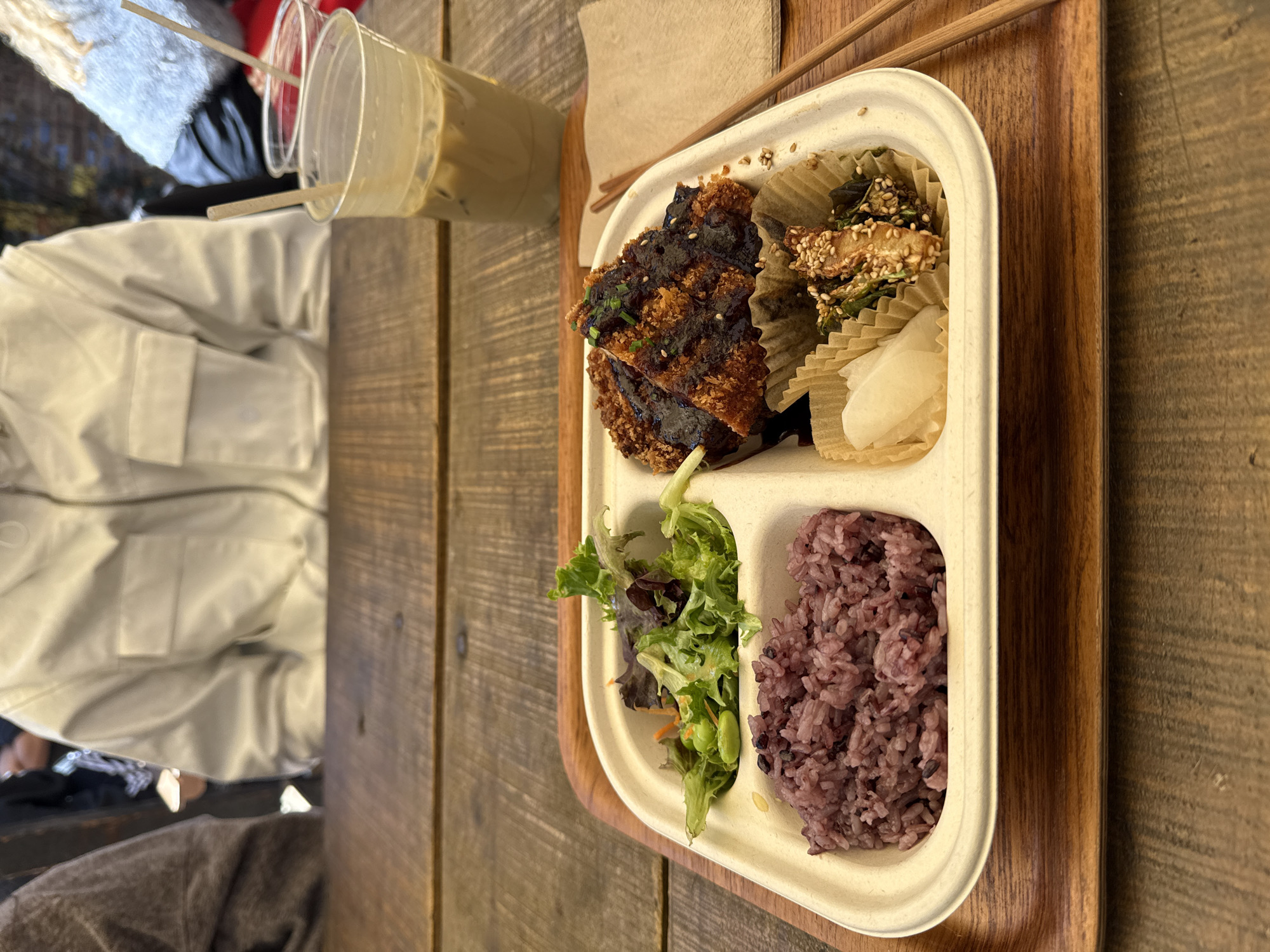Here's a detailed description of the image:
**Scene:** The image shows a meal on a wooden table. The meal is presented in a segmented, rectangular container placed on a wooden tray.
**Meal Components:**
* **Top Left Compartment:** Contains breaded and fried cutlets with a dark, shiny glaze.
* **Top Right Compartment:** Holds small servings in cupcake liners with what looks like vegetables with sesame seeds and small, white pickled radish.
* **Bottom Left Compartment:** Features a small salad with mixed greens, carrots, and what appears to be edamame.
* **Bottom Right Compartment:** Contains purple rice.
**Other Objects:**
* A plastic cup with a light-colored beverage (likely iced coffee) is positioned near the meal.
* A set of chopsticks rests on the edge of the tray.
* A napkin.
* Someone wearing a light-colored jacket is partially visible in the background.
**Location Clues:**
While the image doesn't provide explicit visual clues to determine the location, it is stated that this is in New York City, United States of America. Without landmarks or other contextual information, it's impossible to pinpoint the exact spot.