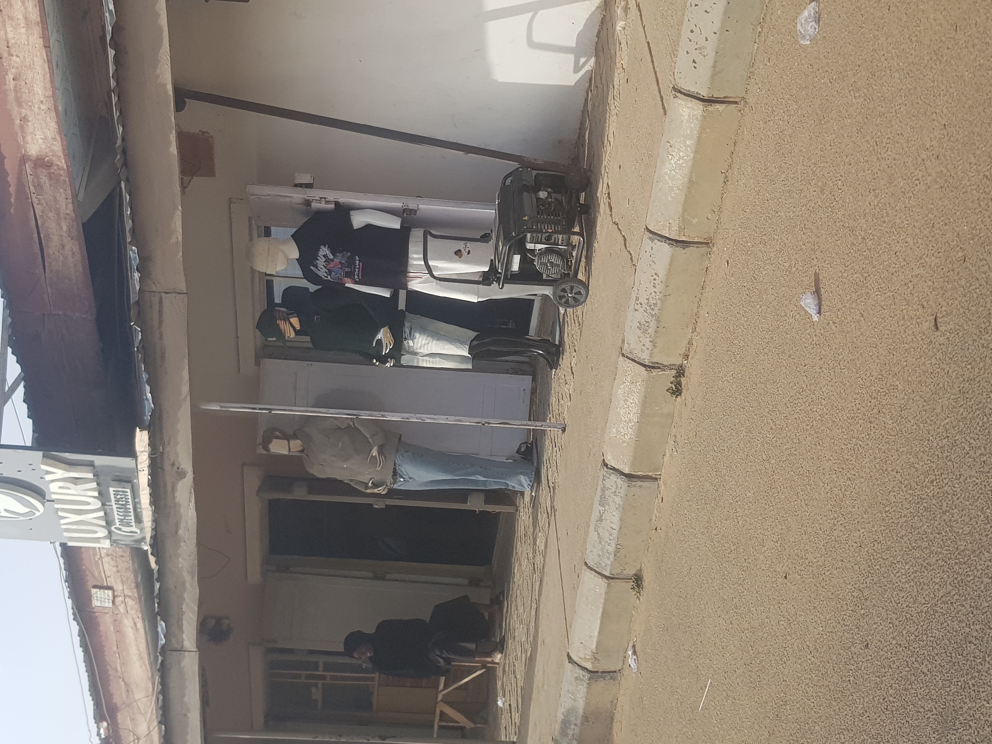 The image shows a storefront in Dabawa, Nigeria. A sign reading "Luxury" is visible. Three mannequins dressed in casual clothing are displayed in the storefront window. A portable generator is situated next to the mannequins. A person is sitting on a stool inside the shop. The street appears to be paved.