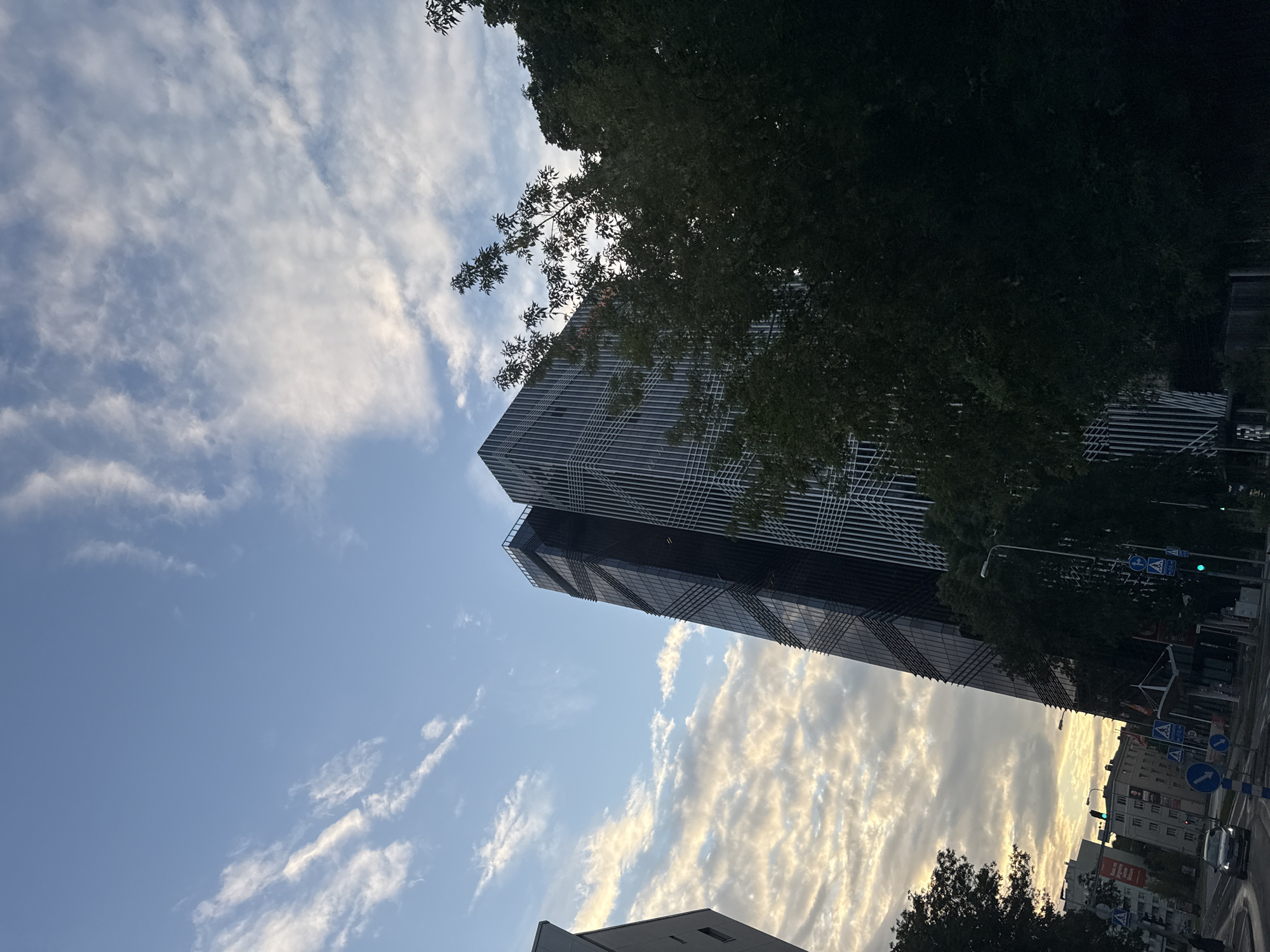 The image shows a modern, dark-glass skyscraper with a distinctive diagonal pattern of reflective louvers, located in Tallinn, Estonia. The building's facade is primarily black glass, with white vertical fins or louvers that create a striped effect. To the left of the skyscraper, another building is partially visible, appearing lighter in color and less ornate. In the foreground, lush green trees obscure parts of the lower sections of the buildings and the street scene. The sky above is a mix of blue sky and scattered white clouds, some of which are illuminated by the setting sun, casting a warm glow. The lower portion of the image reveals a street intersection with traffic lights, road signs, and cars, indicating an urban environment. One car is visible driving on the left side of the road. The scene is set during either sunrise or sunset, judging by the lighting conditions in the sky.