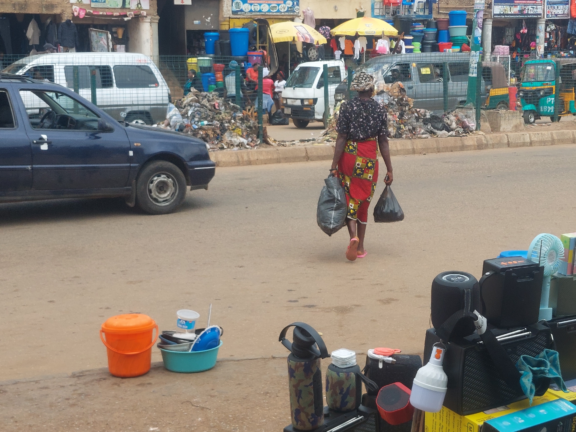 The image depicts a street scene in an urban environment. A woman with a headscarf and a patterned dress is walking away from the camera, carrying two black plastic bags. She is wearing pink sandals. In the foreground, there are several colorful buckets and containers, including an orange bucket and a light blue basin filled with various items. Behind the woman, a busy street is visible with several vehicles, including a blue car in the left foreground, a white van, and a green and yellow motorized tricycle. A large pile of refuse is situated on the side of the road. Shops with awnings and displays of goods line the background. Electronic devices, including speakers and a fan, are displayed on a table in the right foreground. The overall impression is of a bustling market or commercial area.