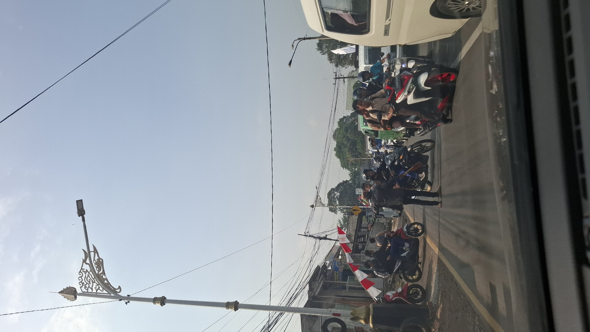 The image captures a street scene in Cilaku, Indonesia, likely near a junction or intersection. The sky is bright and clear, indicating daytime. A prominent street lamp with an ornate design stands on the left side of the road. The road is asphalt with yellow lane markings.
There is a significant amount of traffic, primarily consisting of motorcycles. Several people are visible riding on these motorcycles, some in groups. One man, dressed in a dark jacket and light-colored pants, appears to be directing traffic on foot in the middle of the road. He is facing away from the camera, with his arms extended.
To the left, there are buildings that appear to be shops or residences, with some debris or construction materials visible. Red and white flags, possibly national flags, are displayed on poles near these buildings.
On the right side of the frame, a white van is partially visible, suggesting the presence of other vehicles. The overall impression is of a busy, somewhat congested urban environment in Indonesia.