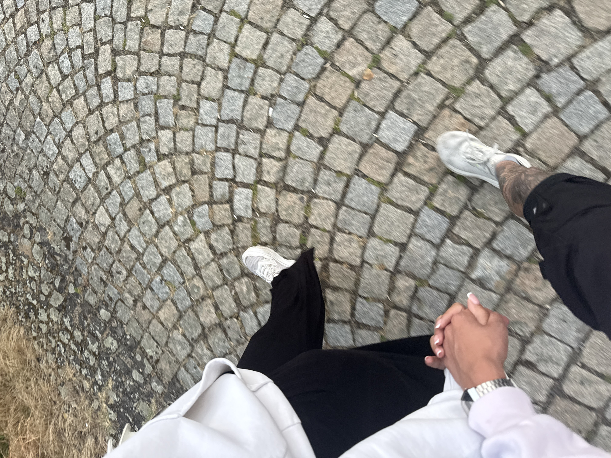 The image shows a close-up of two people's legs and hands as they walk on a cobblestone path. One person is wearing a white hoodie and black pants, with white sneakers. The other person is wearing black shorts and white sneakers with visible tattoos on their leg. Their hands are clasped together. The background is a cobblestone path with some dry grass and dirt to the left. The location is identified as the city of Koprinka, Bulgaria, though the image itself does not contain any explicit visual cues to confirm this specific location beyond the provided metadata. The scene depicts a moment of connection and movement through an outdoor urban or park environment.