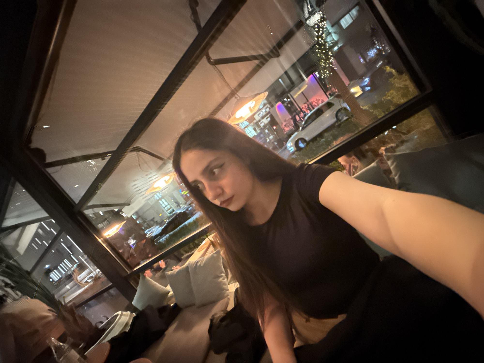 A woman with long, dark hair is sitting on a cushioned seating area inside what appears to be an enclosed outdoor patio. The lighting is dim, with some light sources visible outside the enclosed area. The patio has large windows, offering a view of Yerevan, Armenia, at night, showcasing buildings, streetlights, and a white car parked outside. The woman is wearing a black top and what looks like a black skirt or trousers. She seems to be taking a selfie, with one arm outstretched. Pillows are visible on the couch.